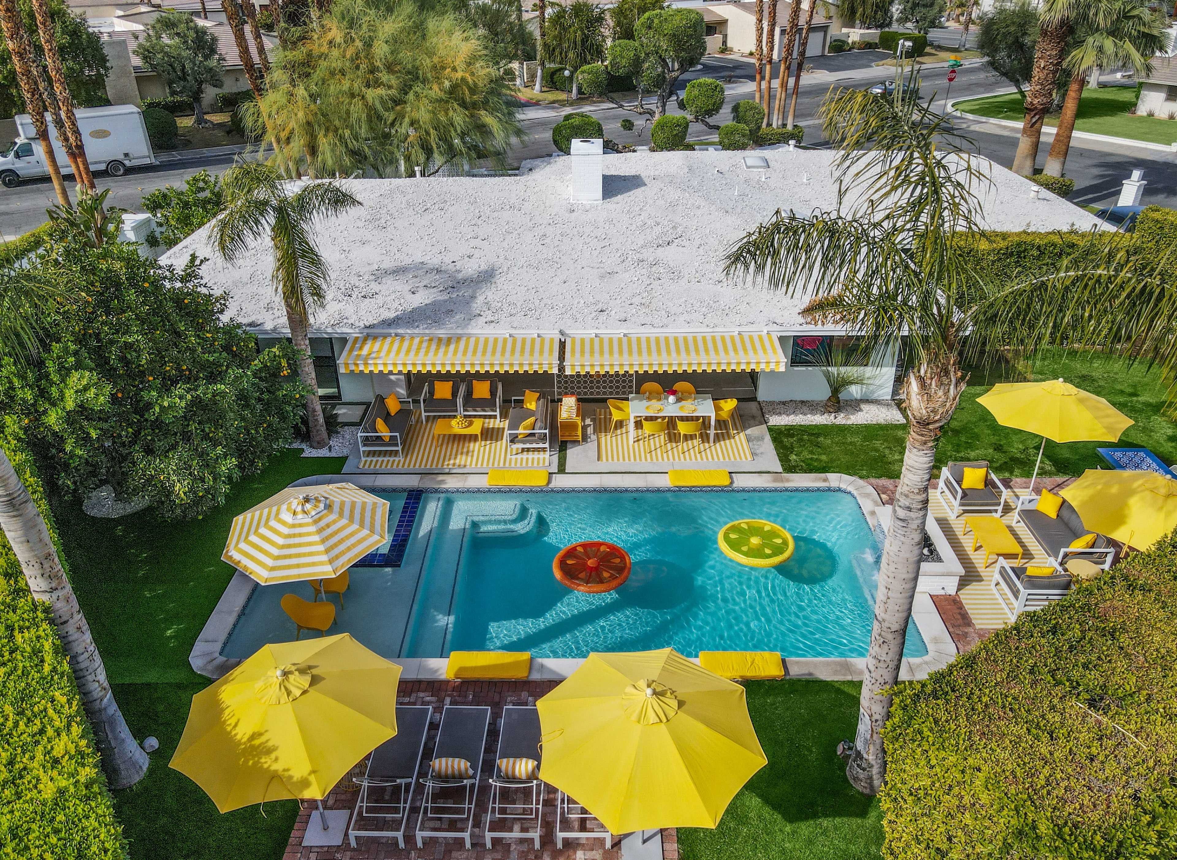 The image shows a backyard pool area with a modern house, featuring yellow umbrellas and lounge chairs around a swimming pool.