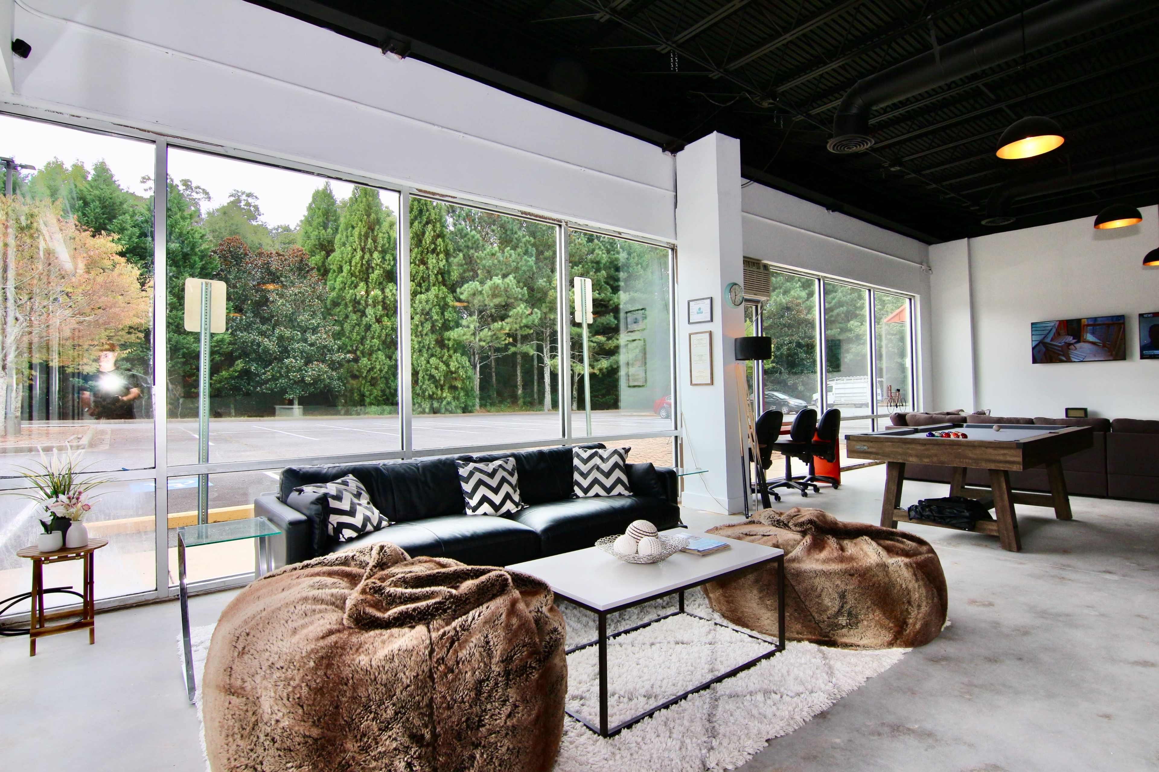 A modern lounge area with a black sofa, a coffee table, and bean bag chairs, surrounded by large windows that overlook a wooded area.