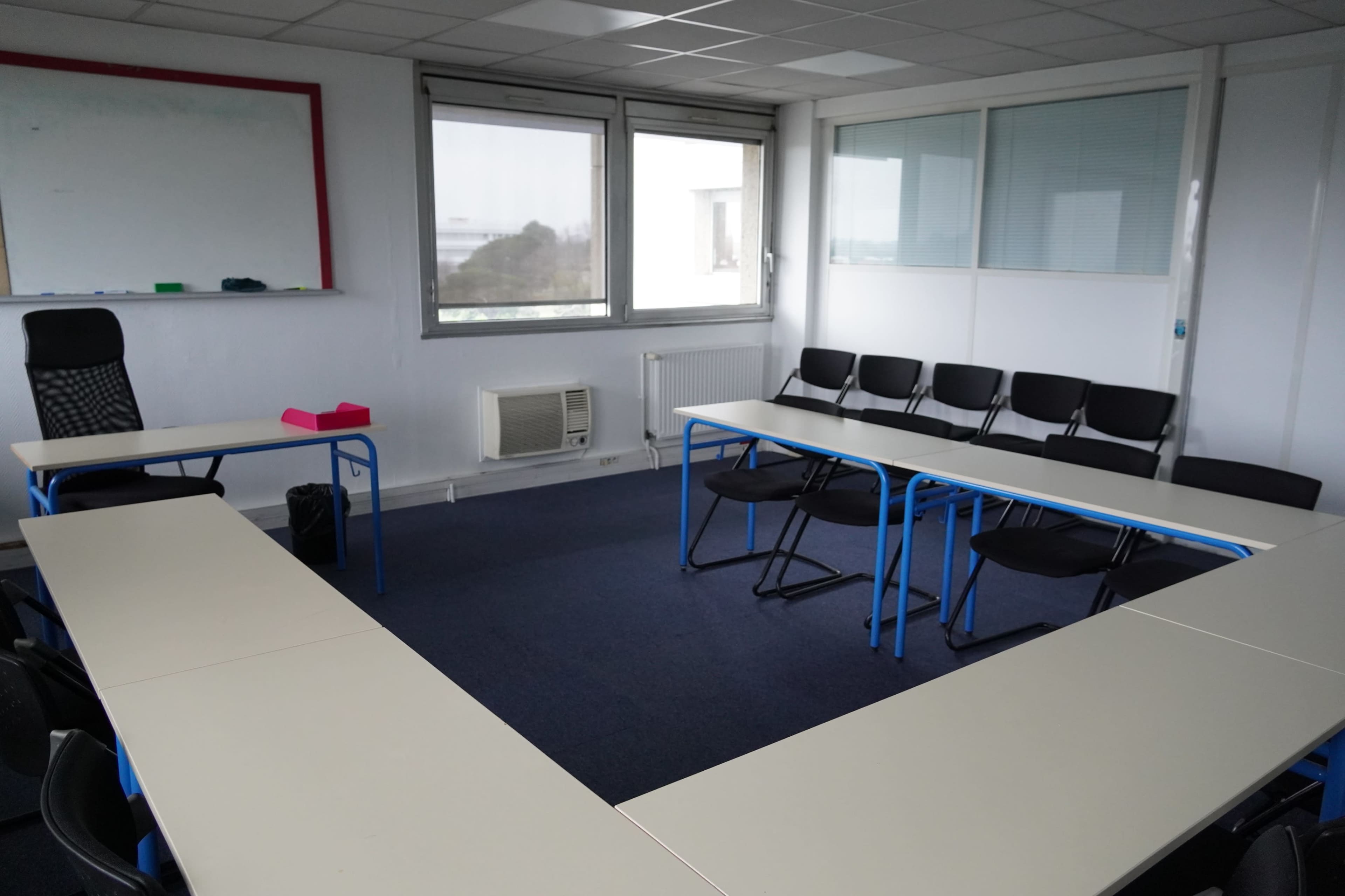 A meeting room with two tables arranged in a U-shape, surrounded by black chairs and equipped with large windows.