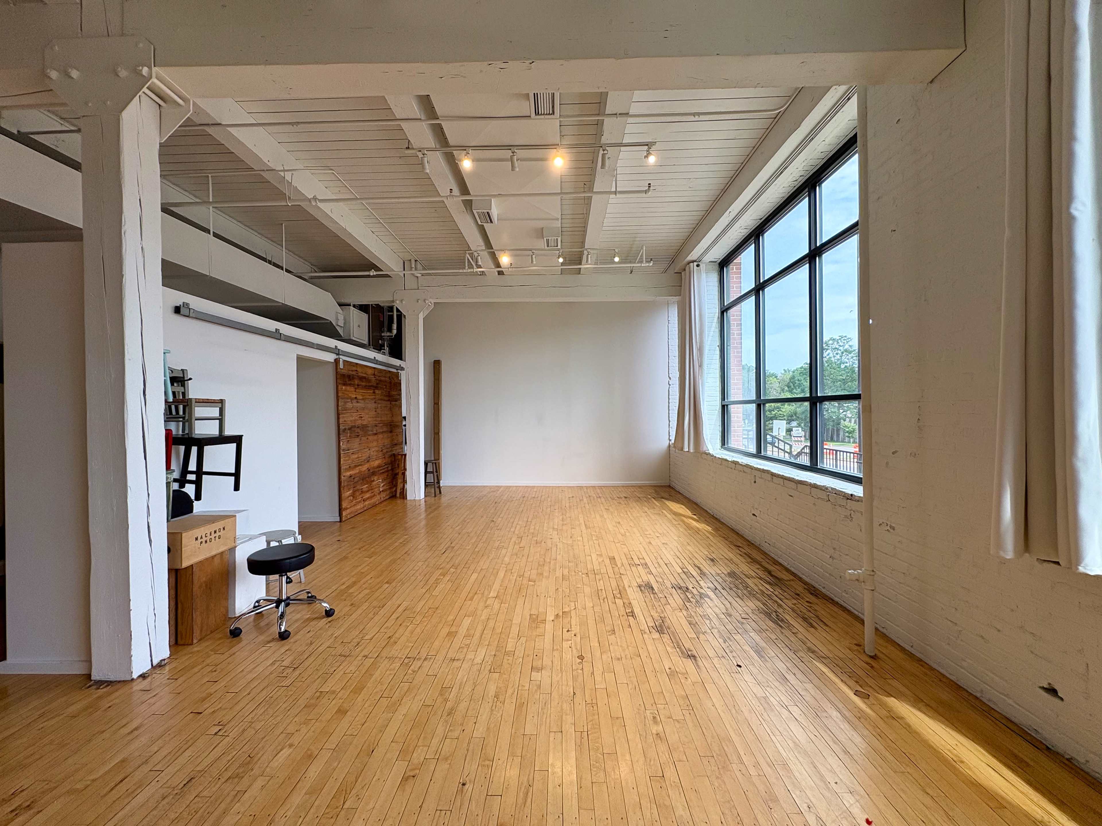 The image shows a spacious, well-lit studio with wooden flooring, large windows, and minimal furnishings.