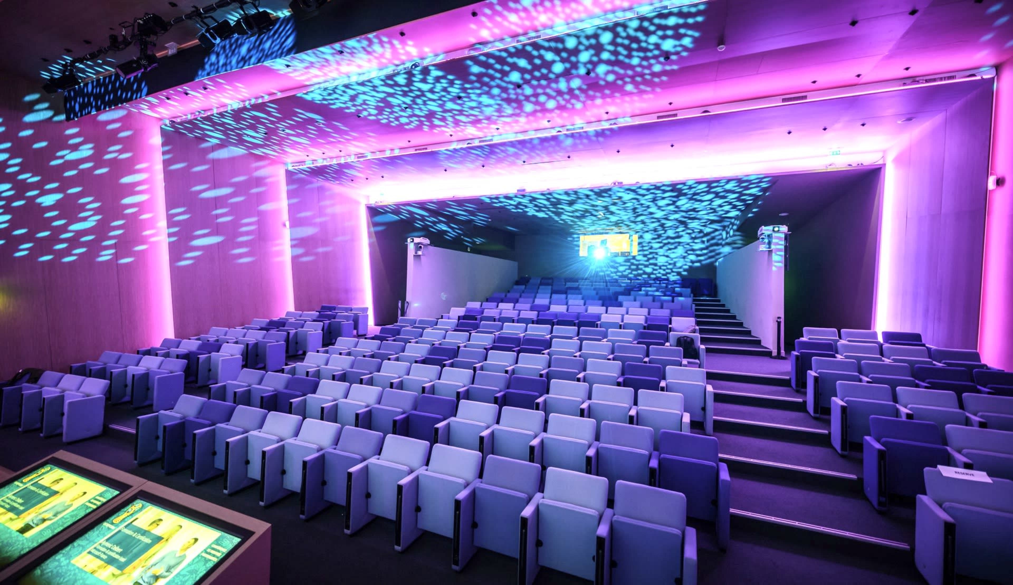 The image shows a modern auditorium with tiered seating and vibrant light projections on the walls and ceiling.
