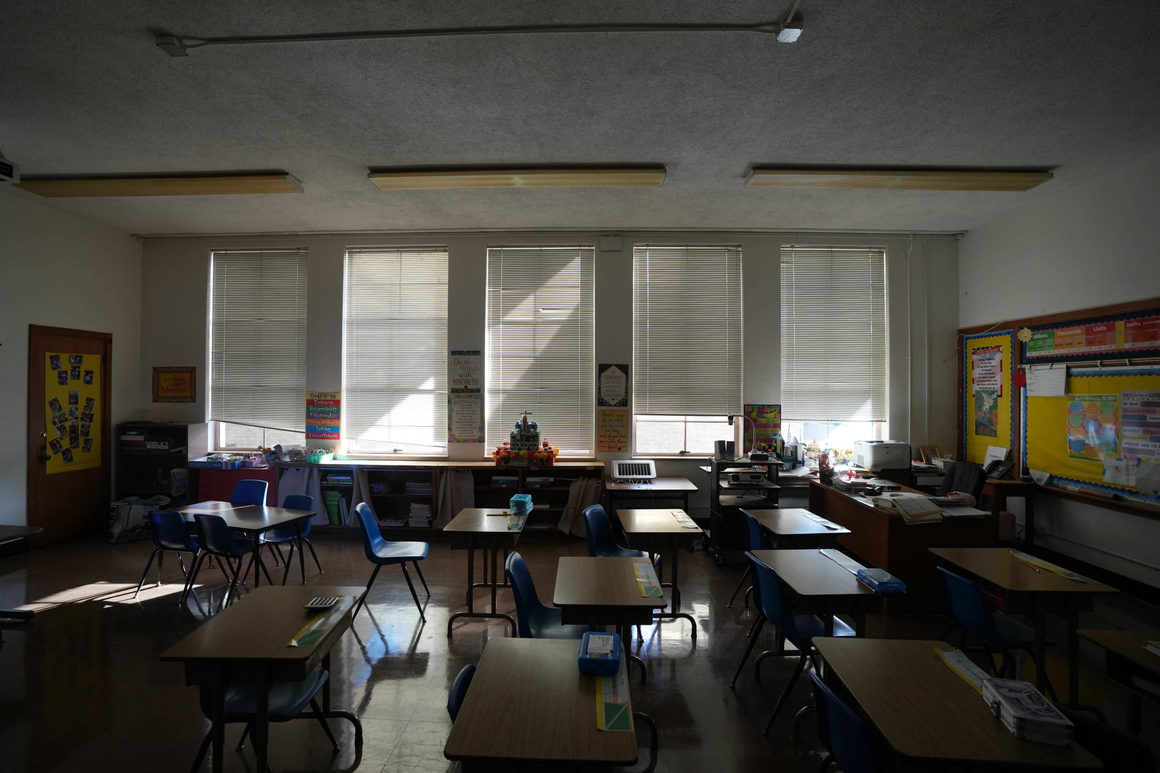 The image shows an empty classroom with desks arranged in rows, natural light coming through the window blinds, and various educational materials on the walls.