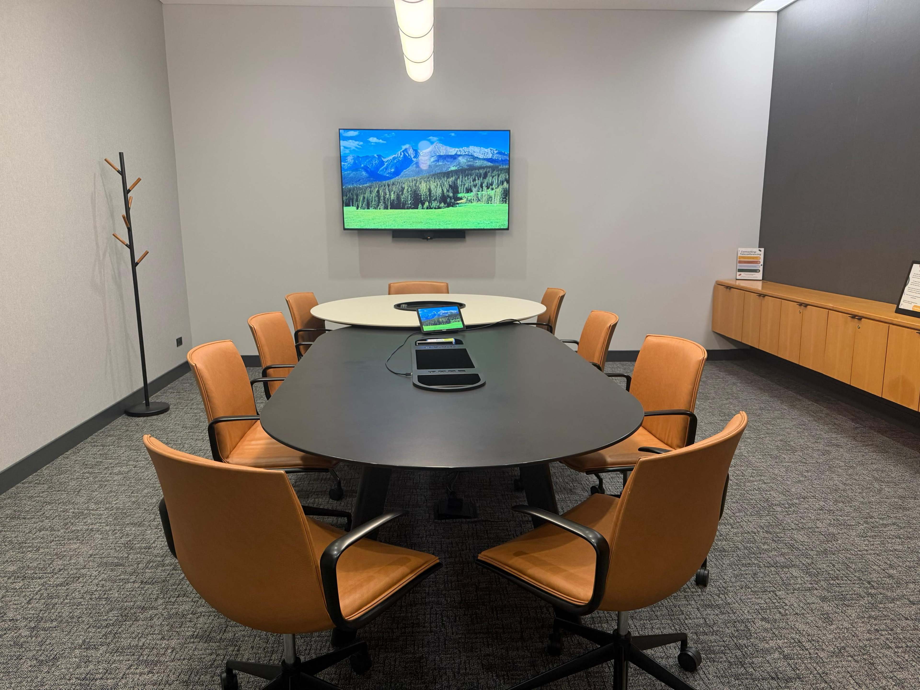 The image shows a modern conference room with a long oval table surrounded by orange chairs, a television mounted on the wall, and a cabinet along the back.