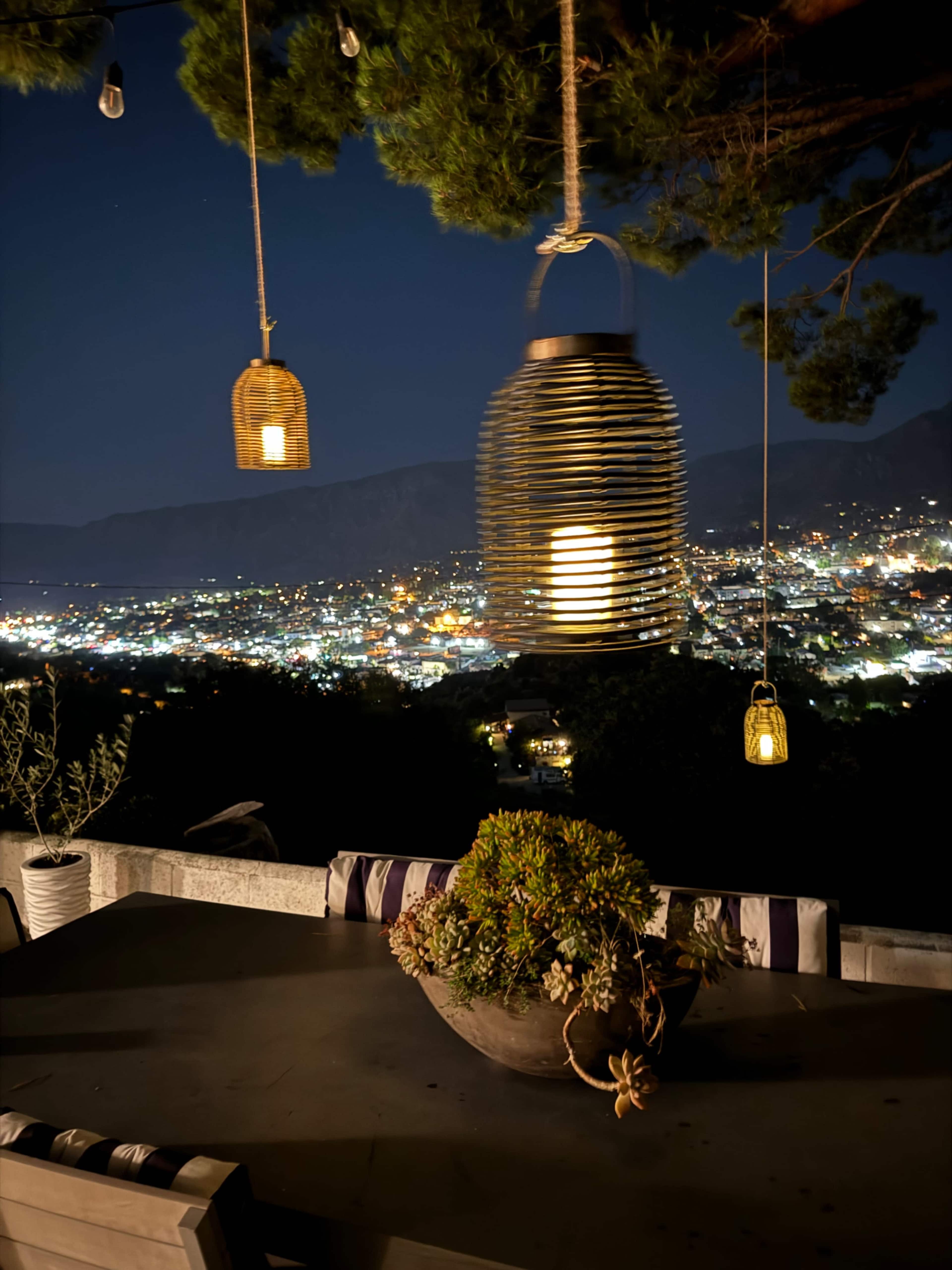 A nighttime view of a city illuminated with lights, framed by hanging lanterns and a table with a bowl of plants.