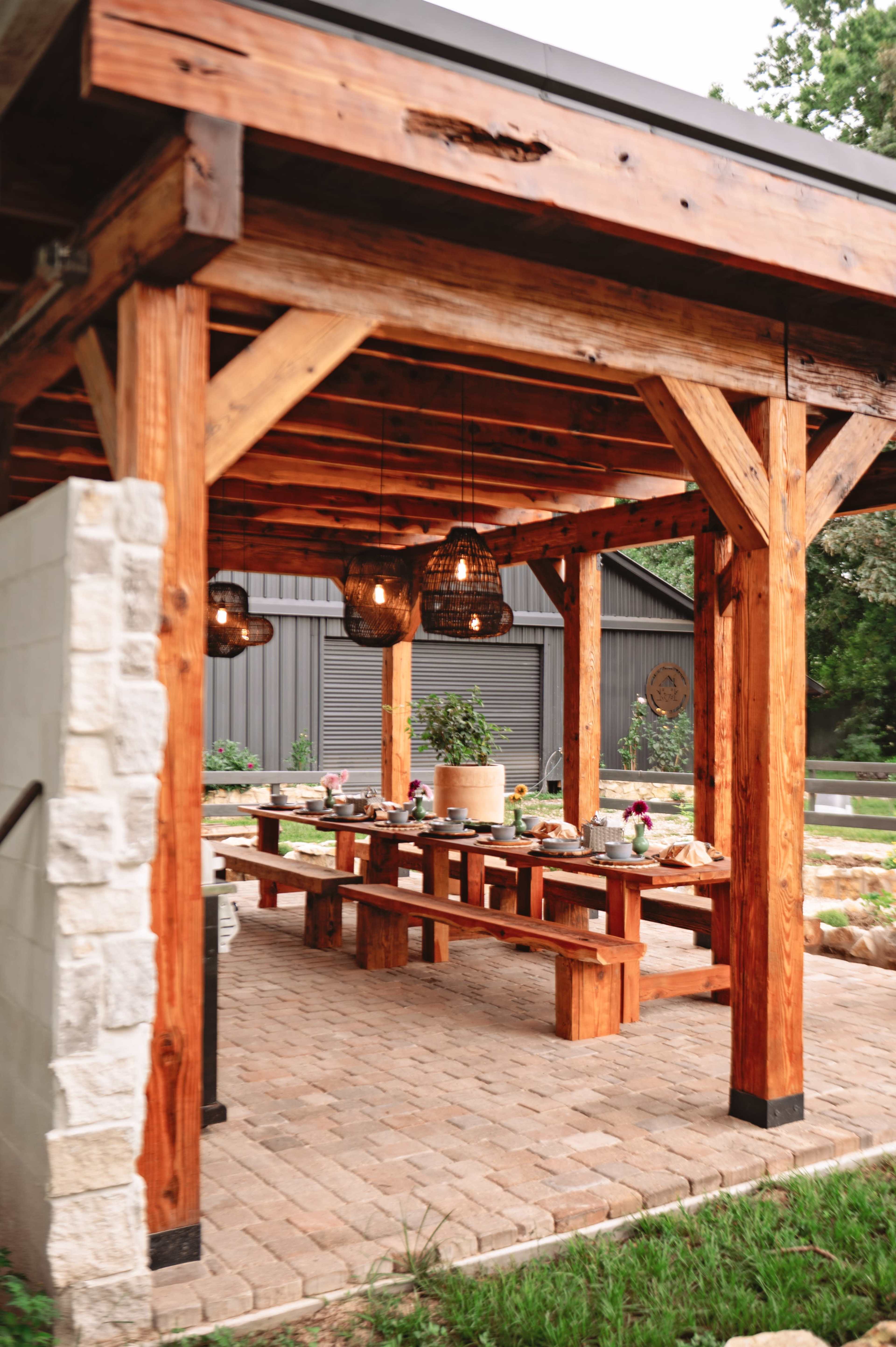 The image shows a wooden pavilion with exposed beams, featuring a long table set for dining, surrounded by hanging light fixtures.