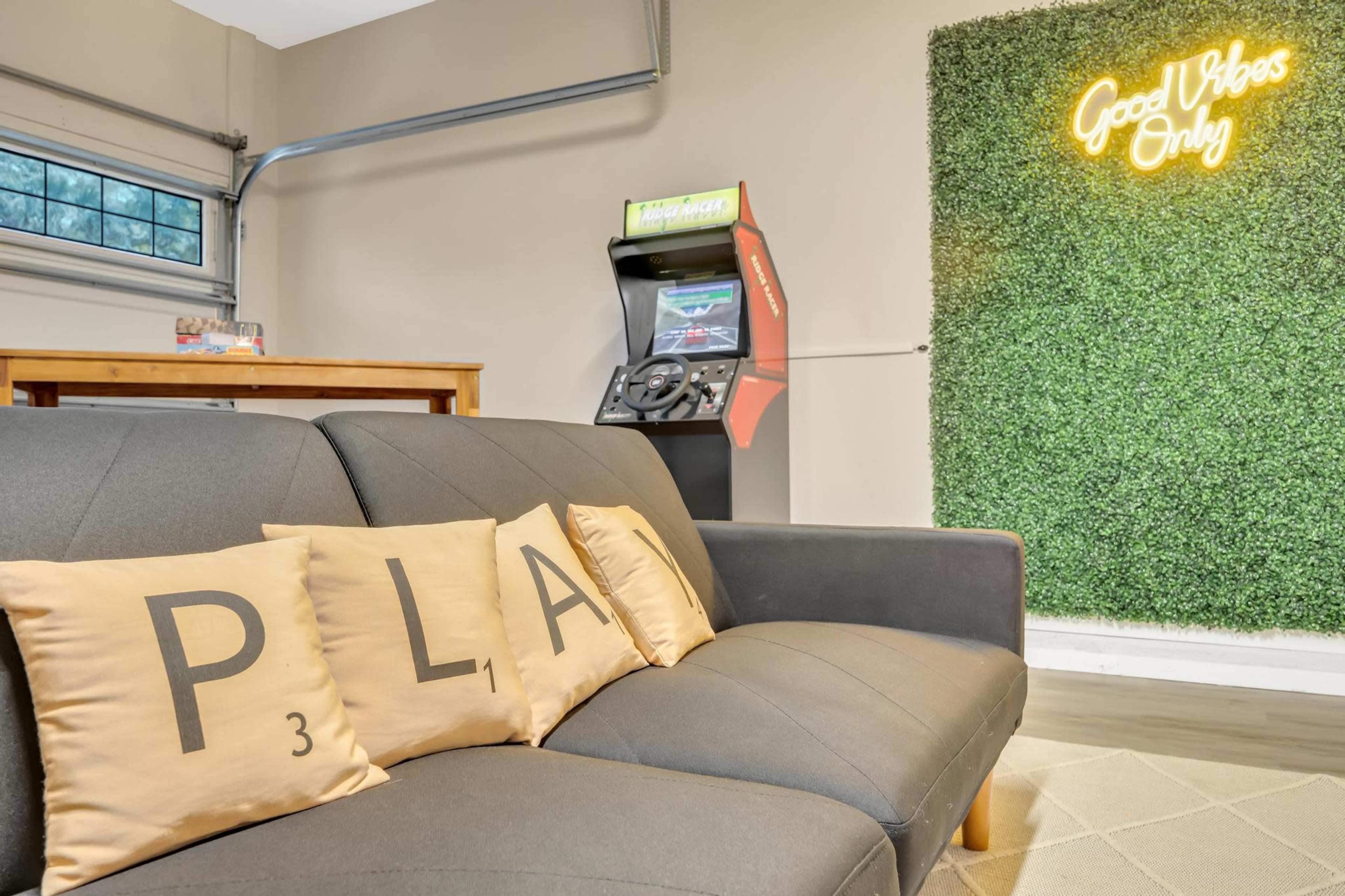 The image shows a cozy room with a couch featuring decorative pillows displaying the letters "P," "L," "A," and "3," alongside a retro arcade game and a neon sign that reads "Good Vibes Only" on a green wall.