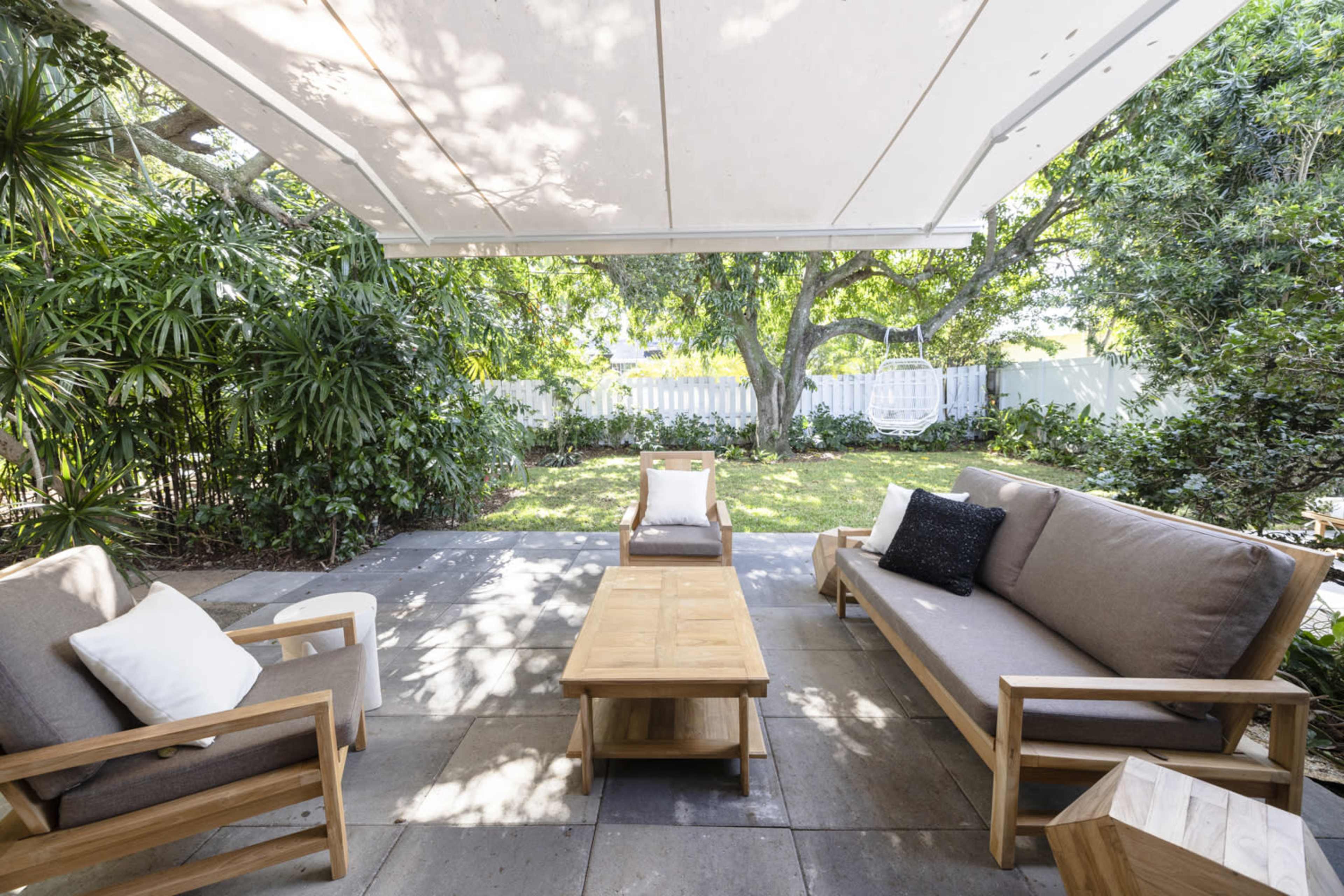 A covered patio features wooden furniture, including a coffee table and two chairs, surrounded by greenery and a lawn.