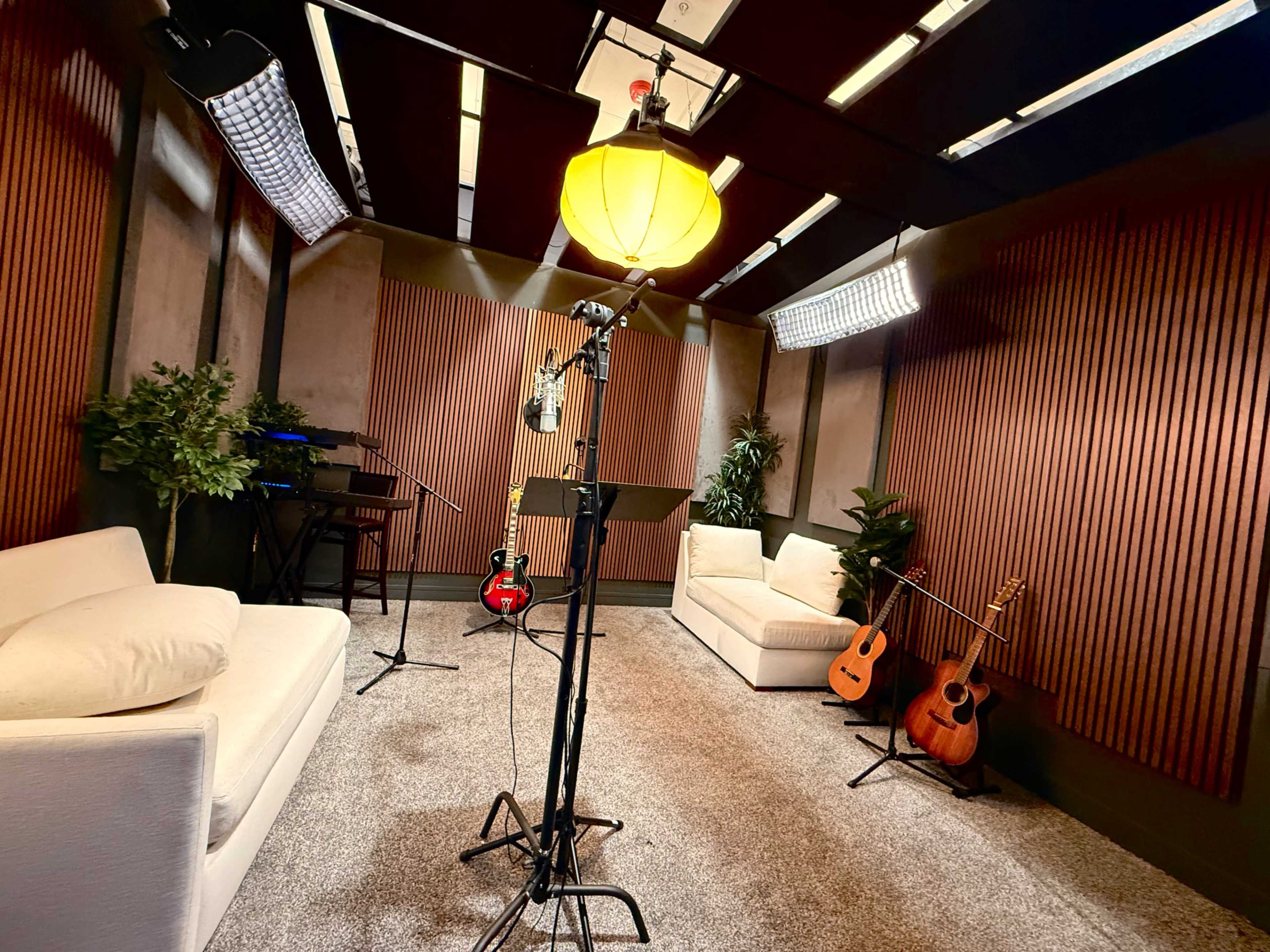 The image shows a sound studio with two white sofas, musical instruments including guitars, and acoustic panels on the walls, along with microphones and lights set up for recording.