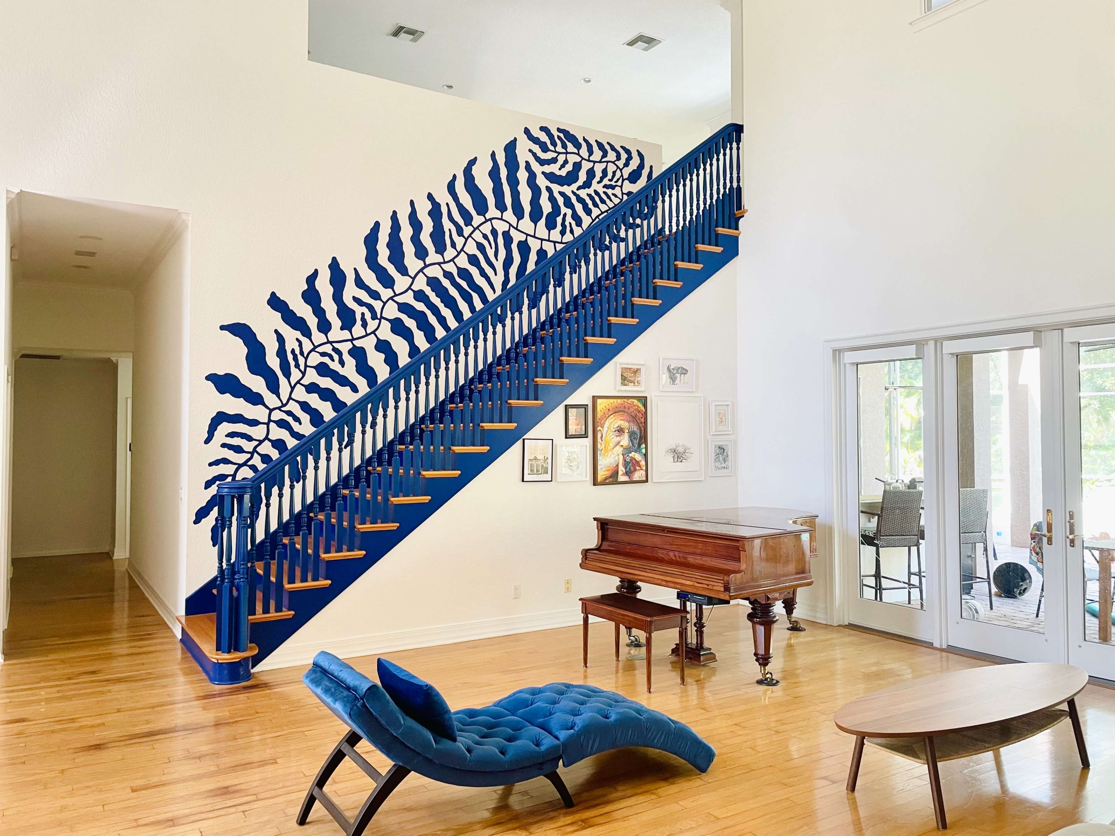 The image shows a bright, modern interior featuring a blue staircase with a patterned wall mural, a grand piano, and a stylish seating area.