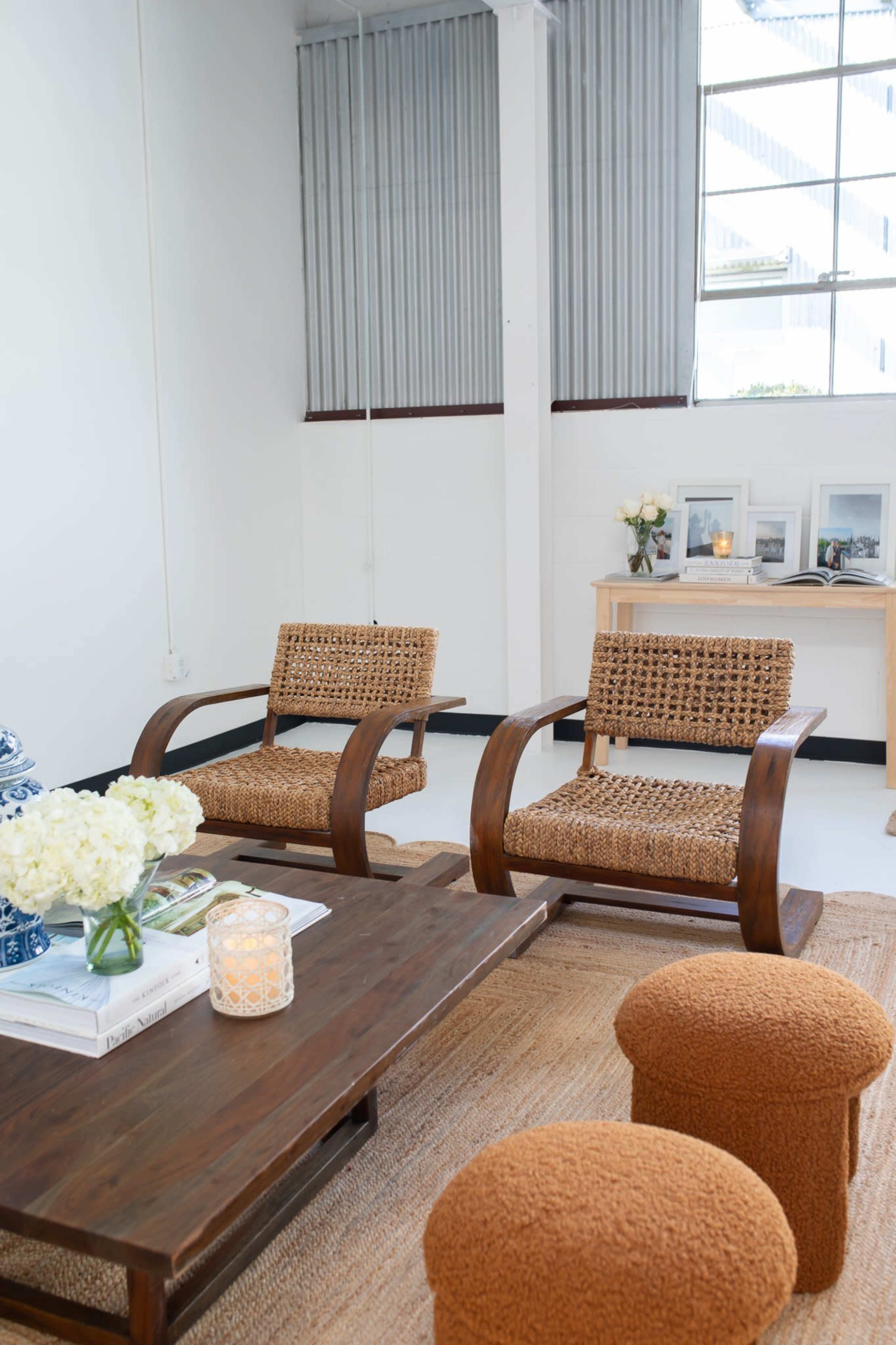 The image shows a minimalist living space featuring two woven armchairs, a wooden coffee table, and decorative elements like a vase with flowers and framed pictures.