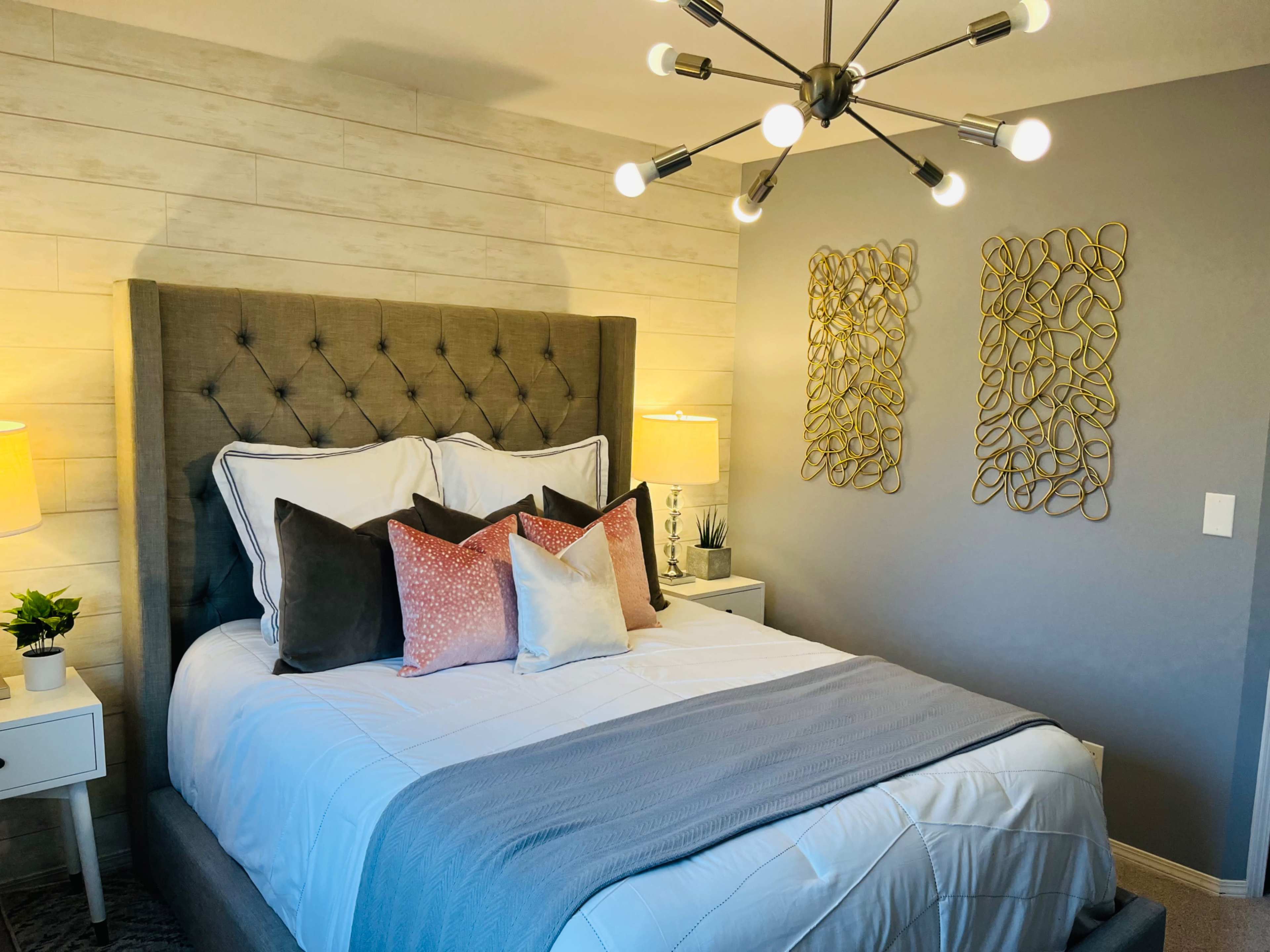 A neatly arranged bedroom featuring a tufted gray headboard bed with decorative pillows, two bedside lamps, and modern wall art.
