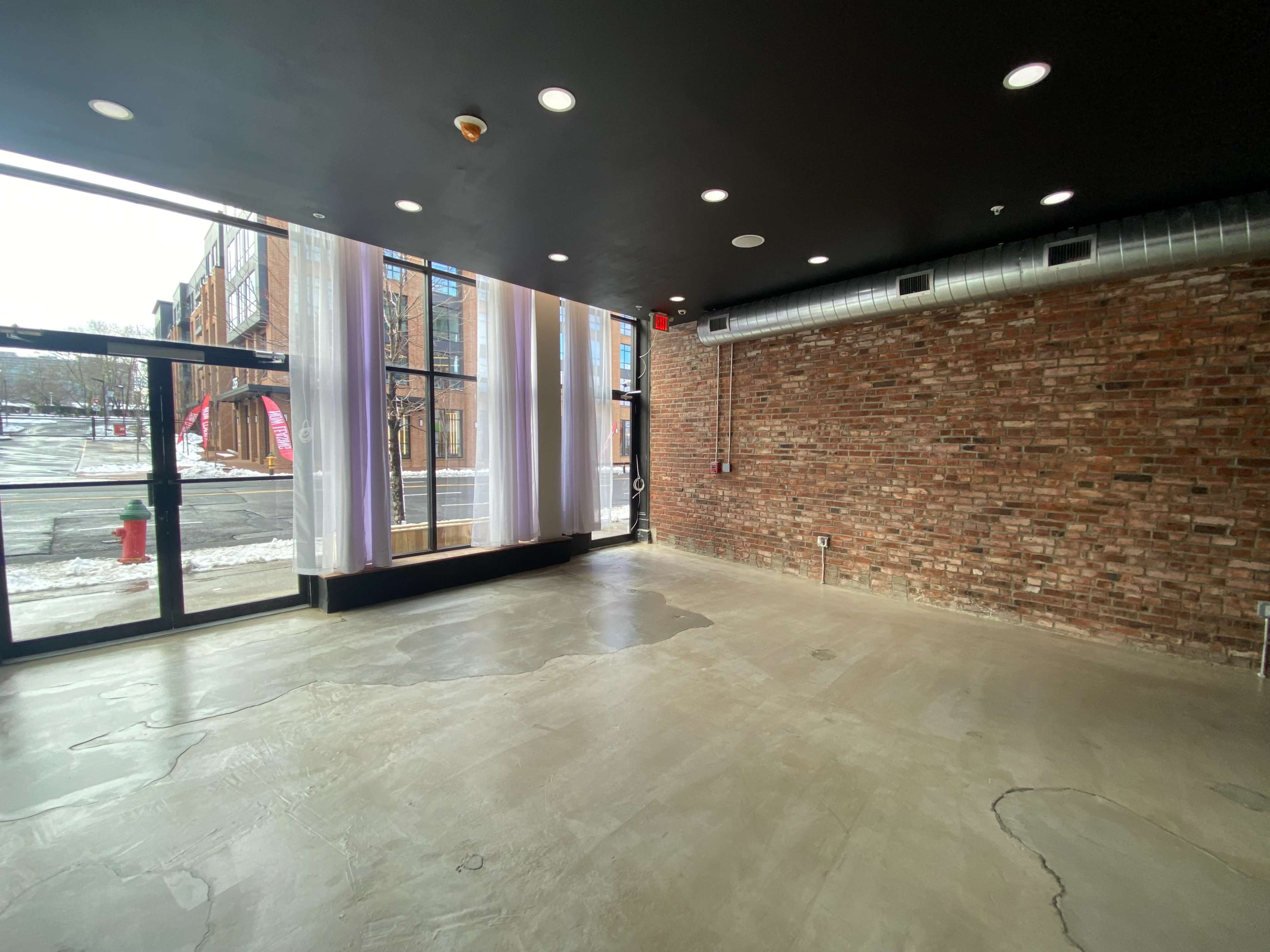 The image shows a vacant commercial space with exposed brick walls, large windows, and a concrete floor.