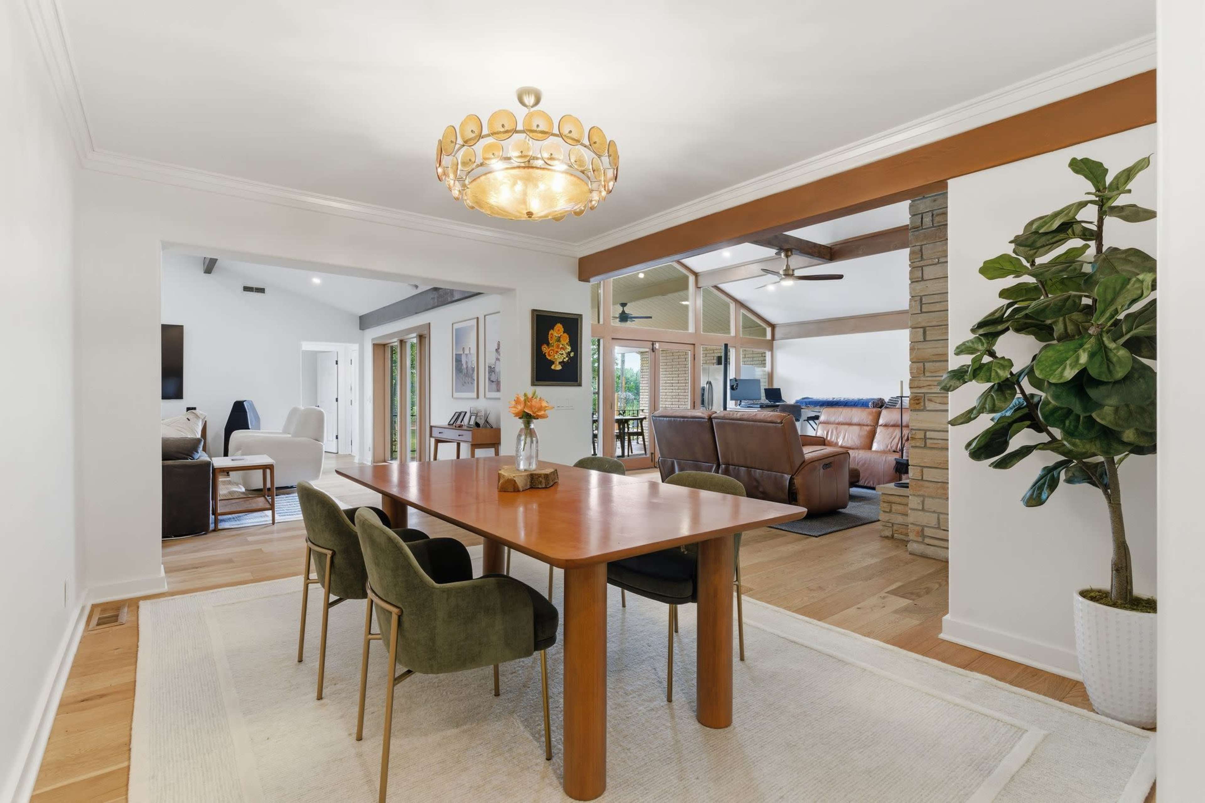 The image shows a dining area featuring a wooden table with four chairs, a chandelier above, and an open view into a living space with comfortable seating and a large window.