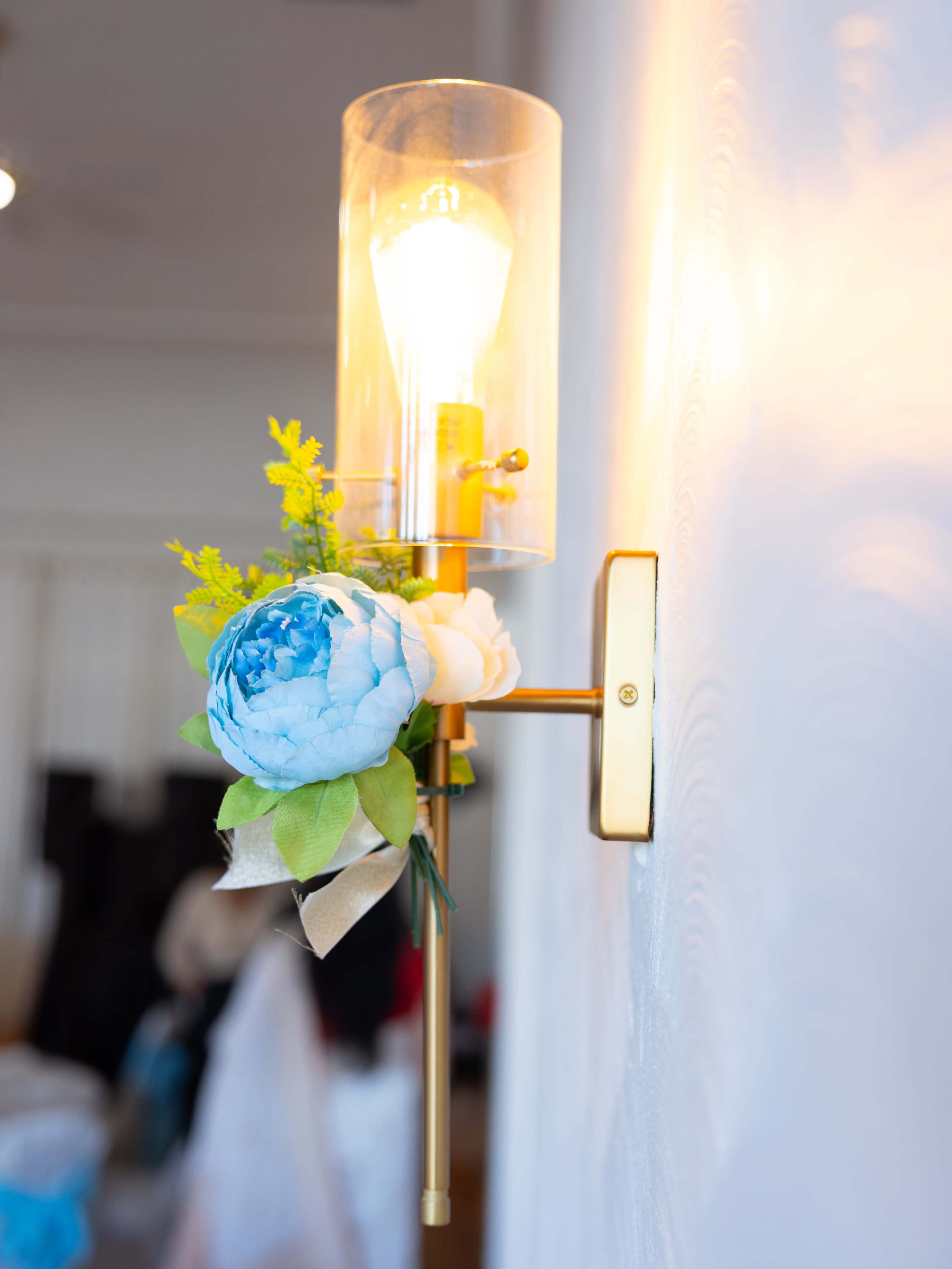 A wall-mounted light fixture with a clear glass shade and flowers is illuminated by a warm light.
