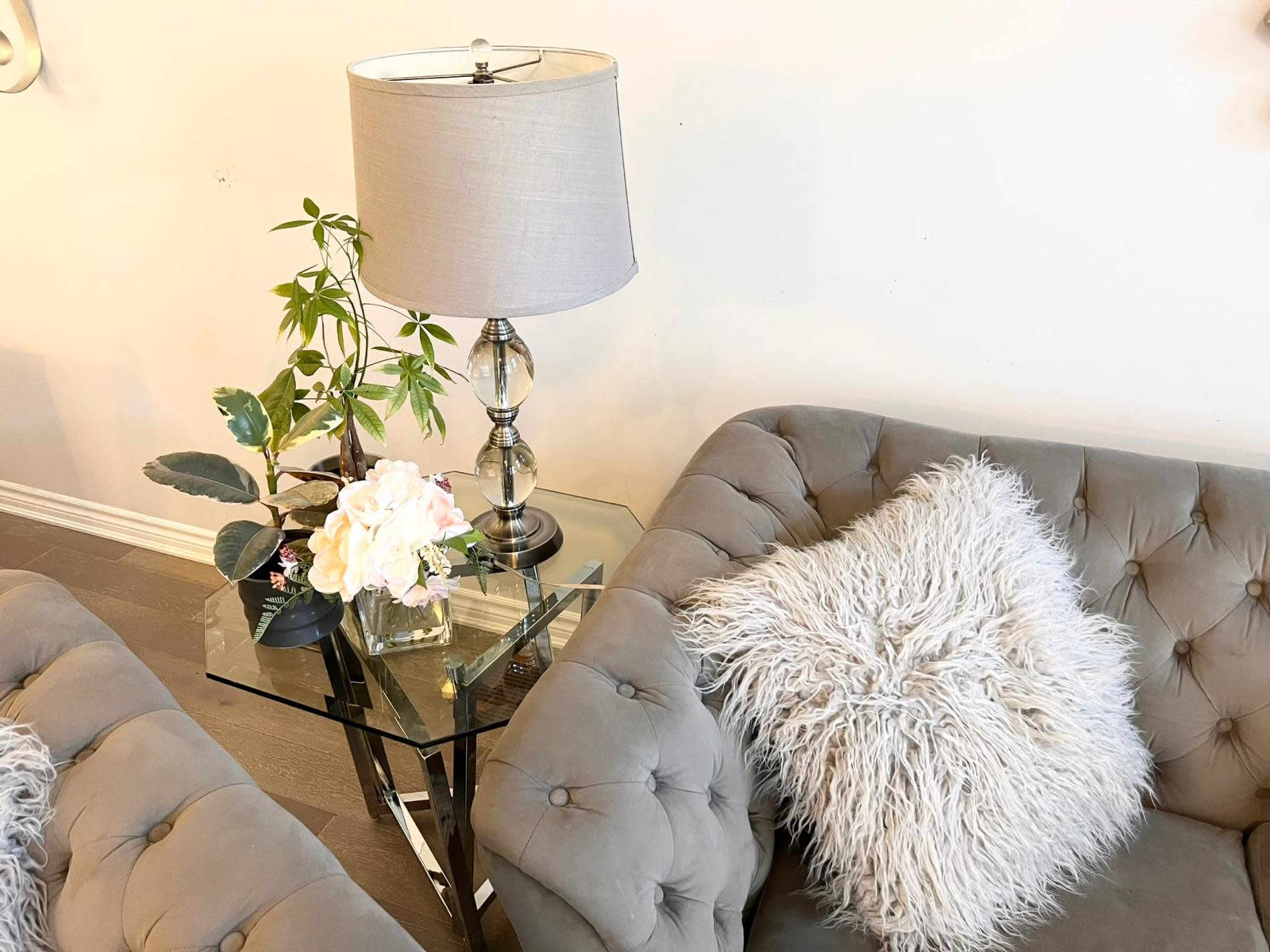 A glass side table with a lamp and a floral arrangement sits between two tufted gray sofas adorned with fluffy pillows.