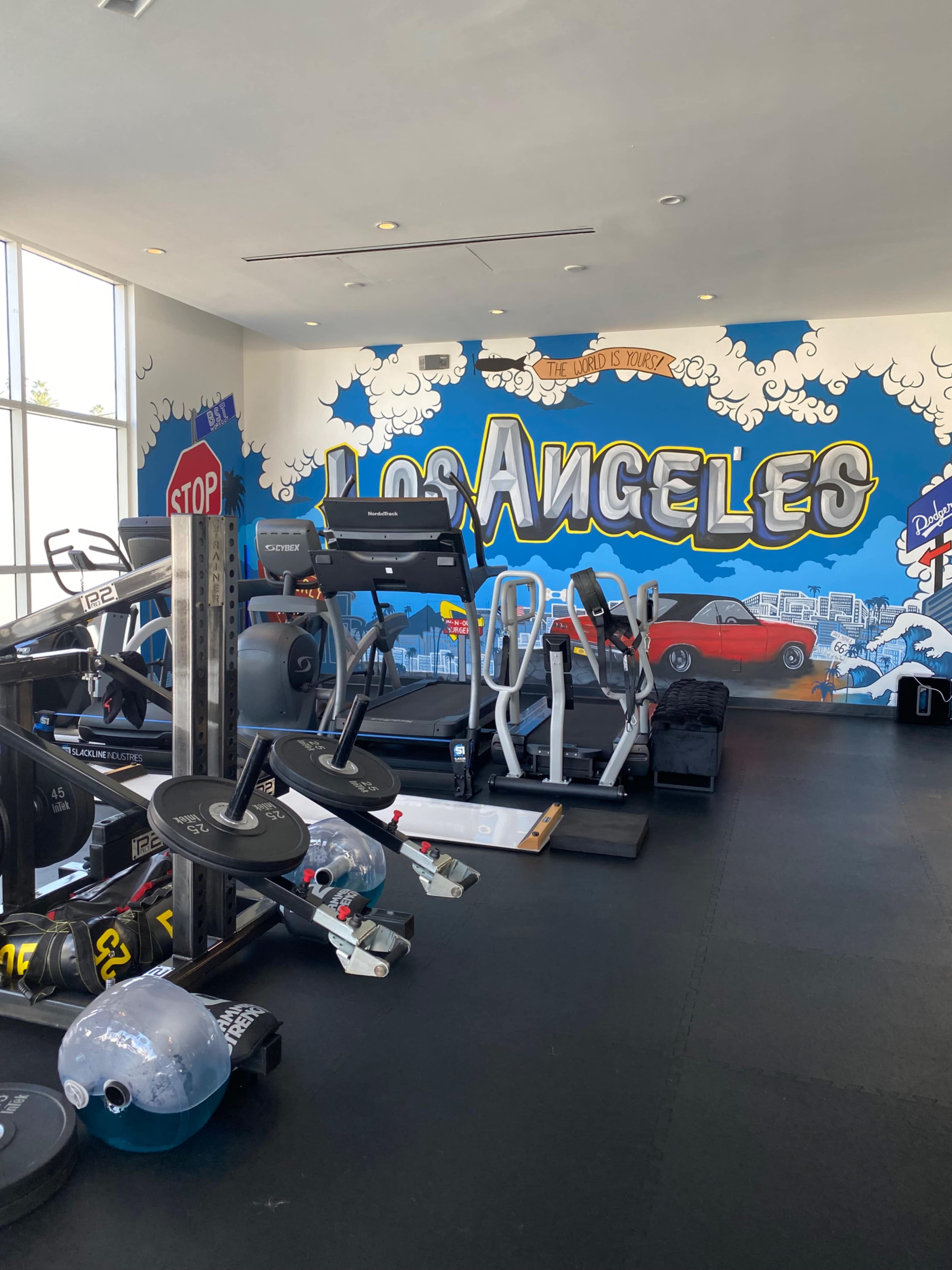 The image shows a gym interior featuring various workout equipment and a colorful mural with "Los Angeles" painted on the wall.