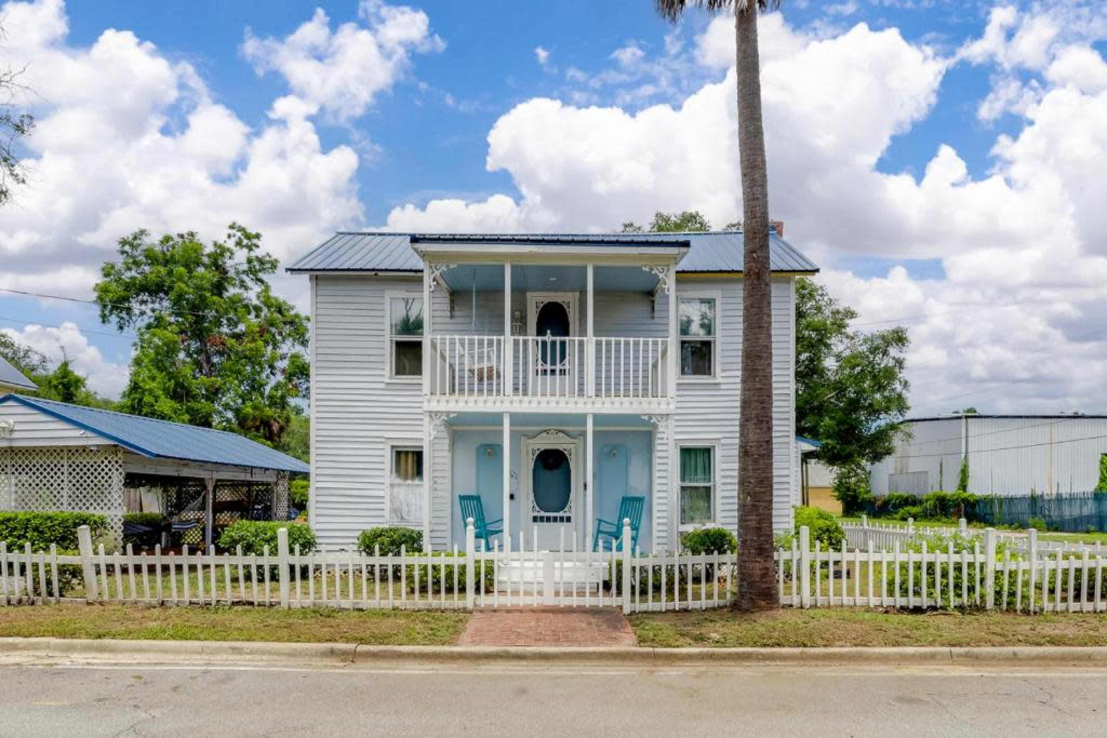 A two-story, light blue house with a white picket fence and a front porch featuring two chairs is situated along a road under a partly cloudy sky.