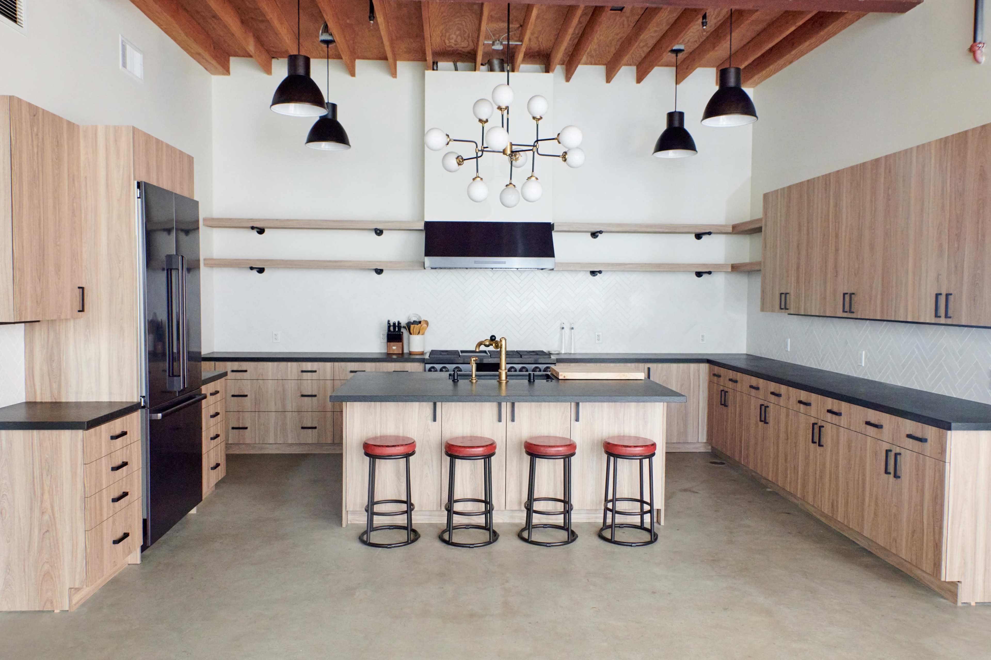 The image shows a modern kitchen with wooden cabinetry, black countertops, and four red-stool seating at an island.