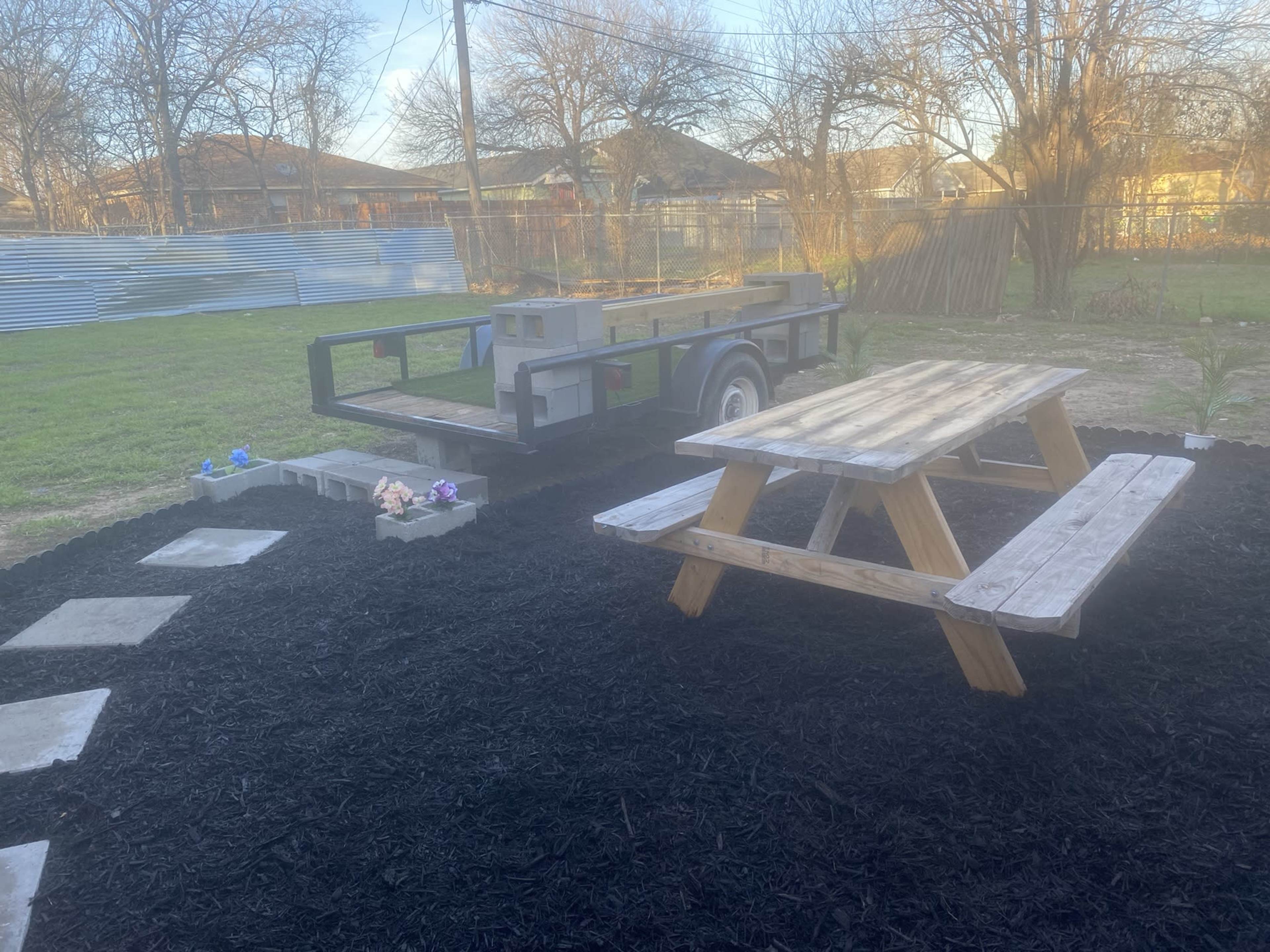 A picnic table is placed on black mulch next to a trailer in a grassy yard.