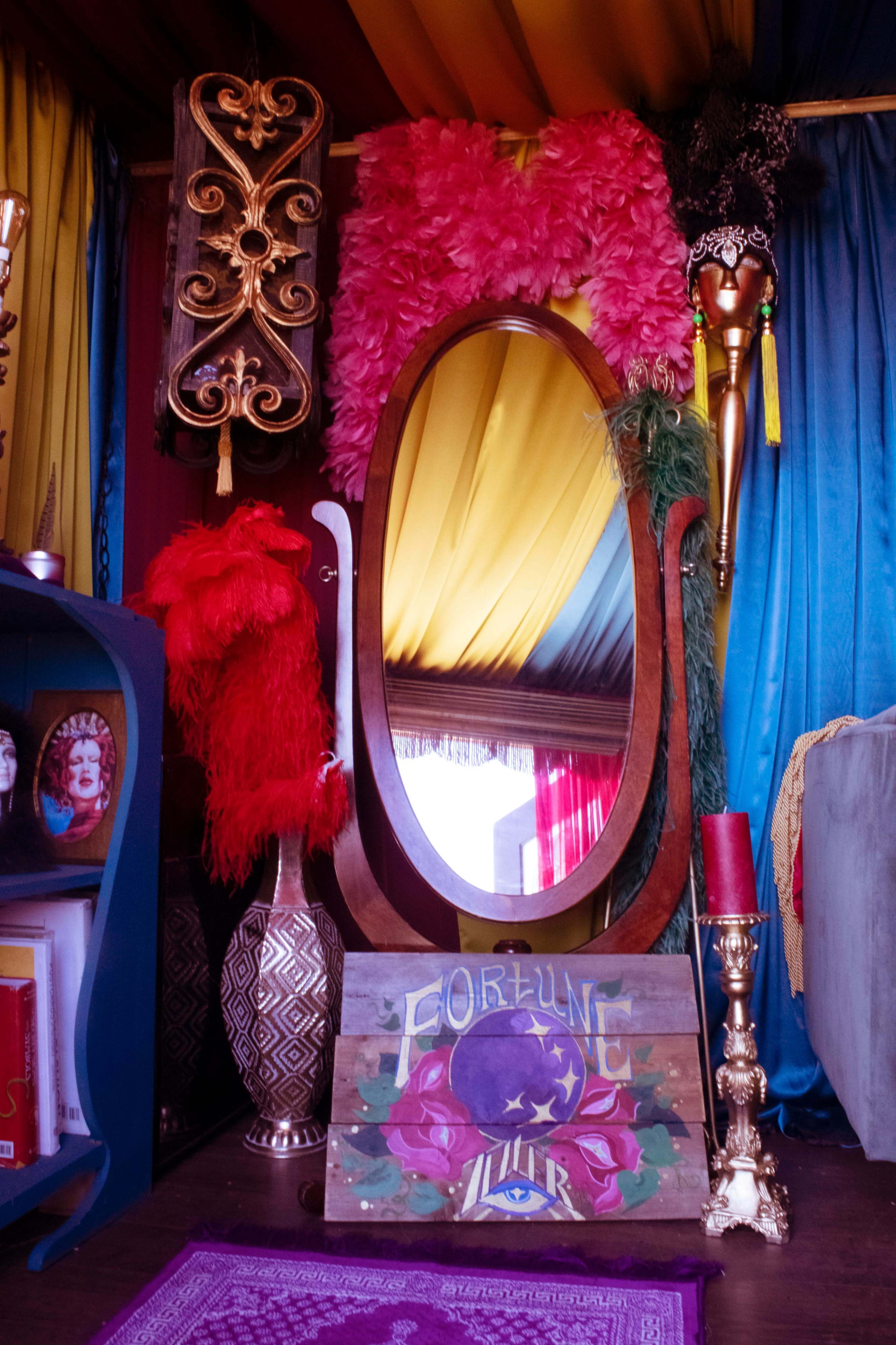 The image features a colorful setting with a large mirror framed in wood, decorated with feathers and a sign that reads "FORTUNE TELLER."