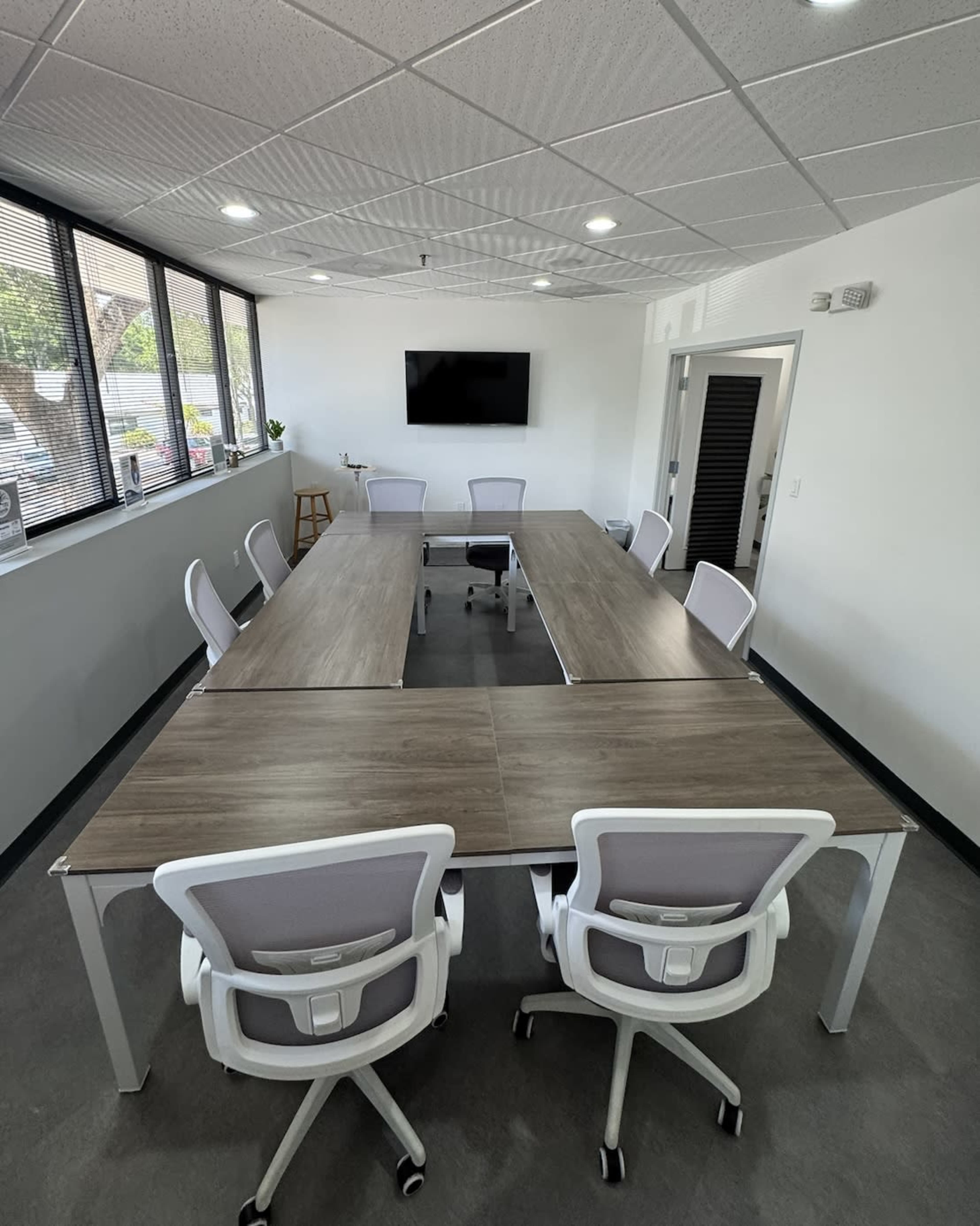 A long conference table with eight ergonomic chairs surrounds it in a modern meeting room with large windows and a wall-mounted screen.