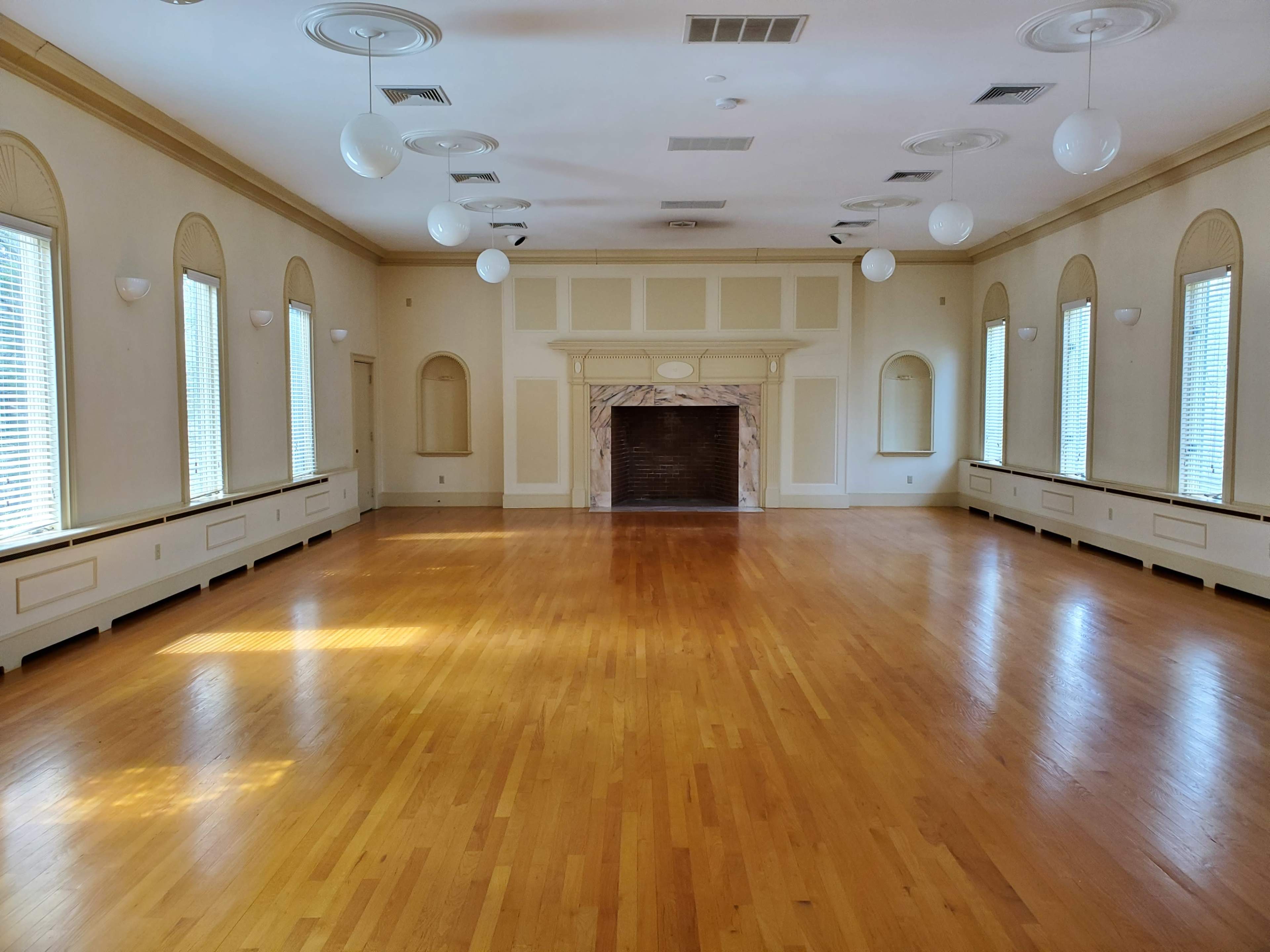 An empty room with wooden floors, large windows, and a decorative fireplace at one end.