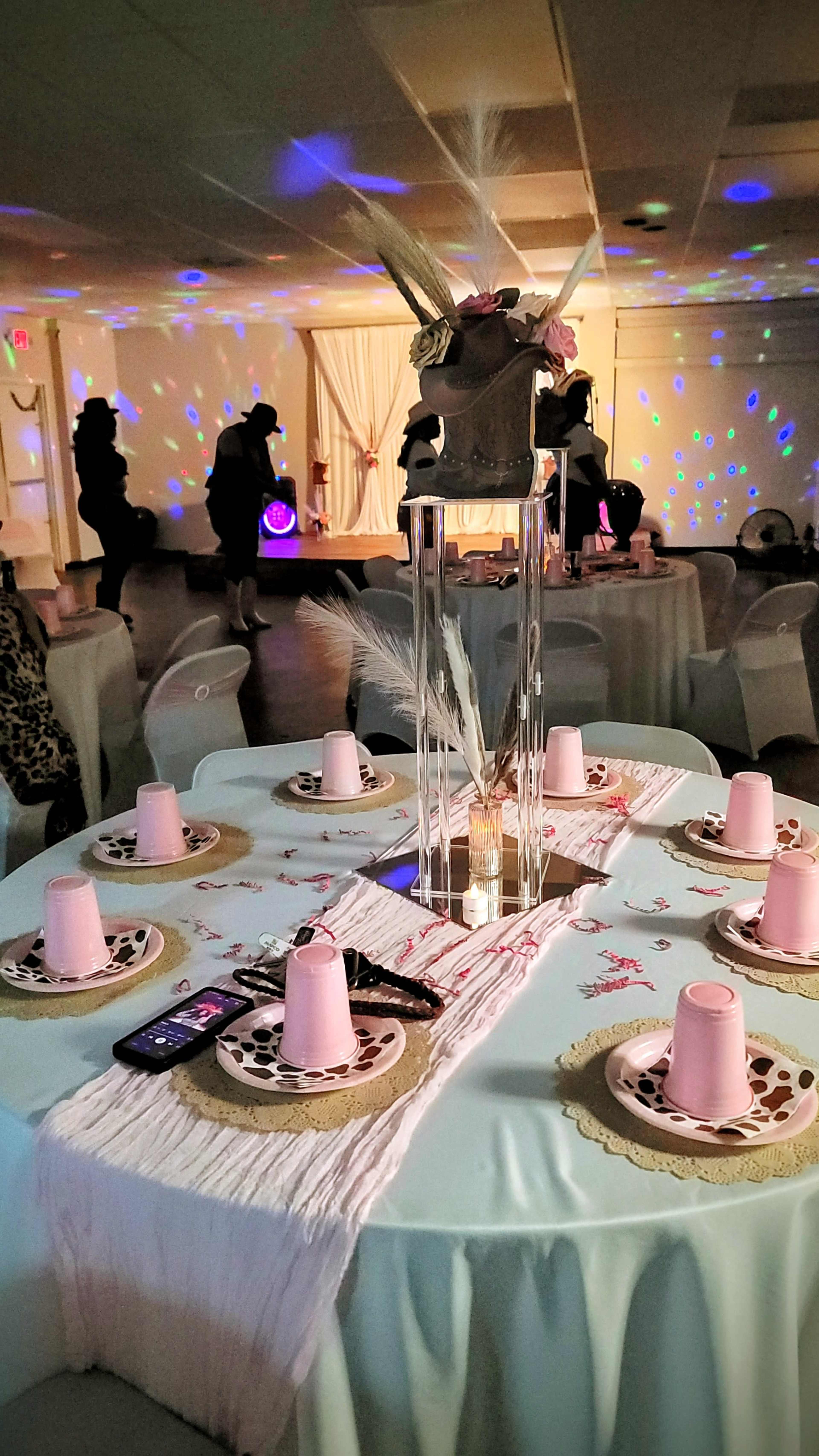 A decorated table is set with pink cups, plates, and a central floral arrangement under soft lighting, while guests are blurred in the background.