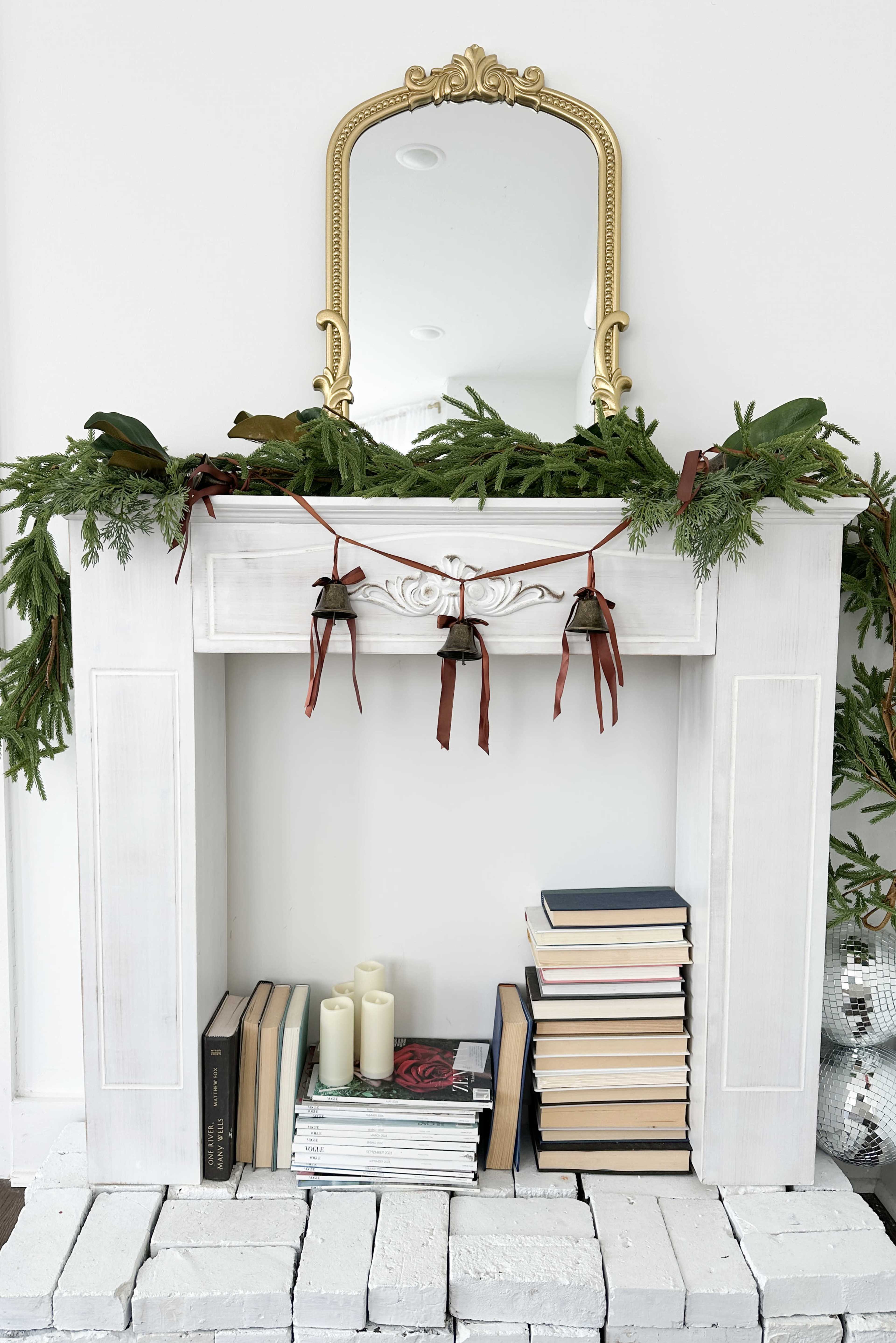 A white mantel with festive greenery and tied bells is decorated with a mirror above and stacked books below.