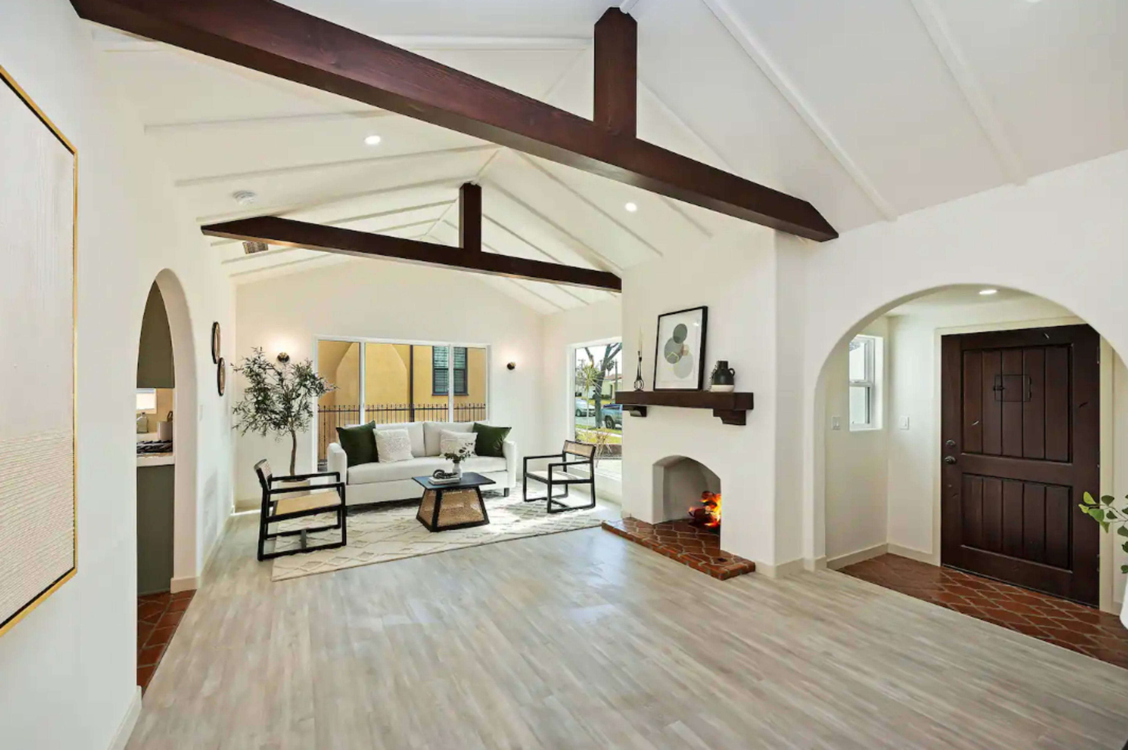A spacious living room with a high-beamed ceiling, light wood flooring, and a seating area featuring a sofa and coffee table next to a decorative fireplace.