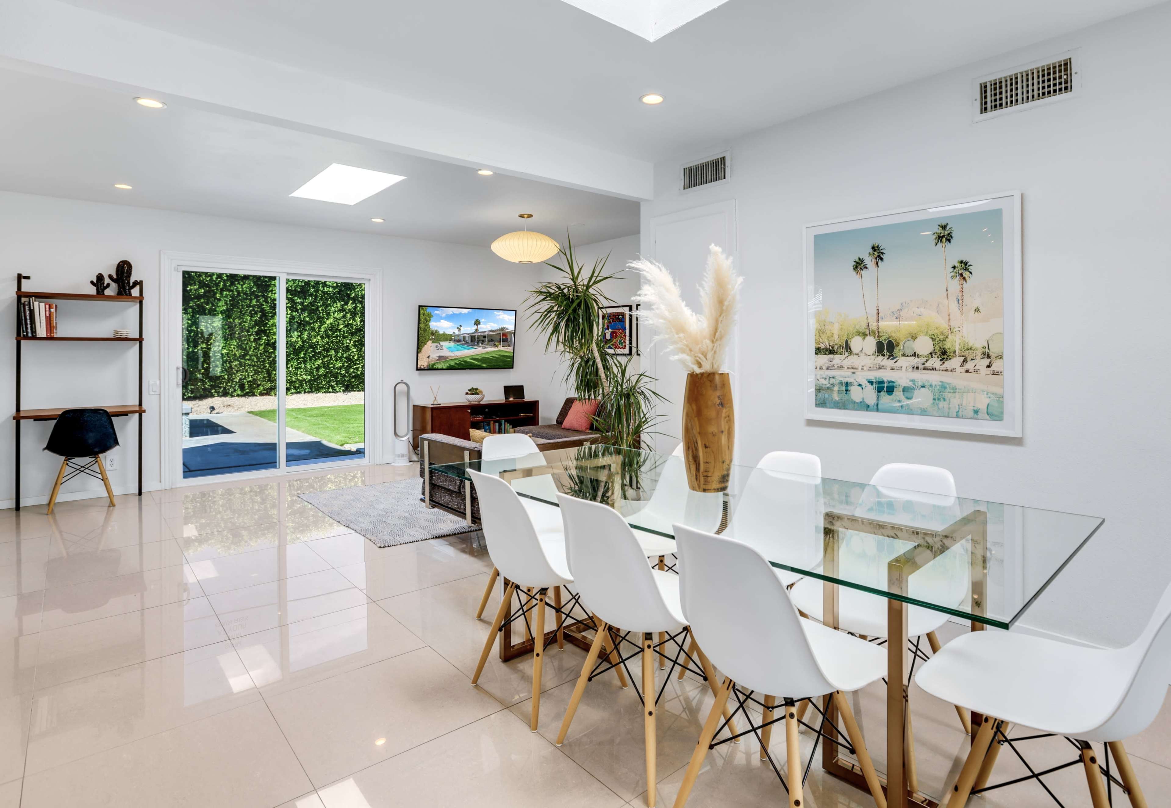 A modern dining area features a glass table surrounded by white chairs, with sliding doors that open to a pool area visible in the background.