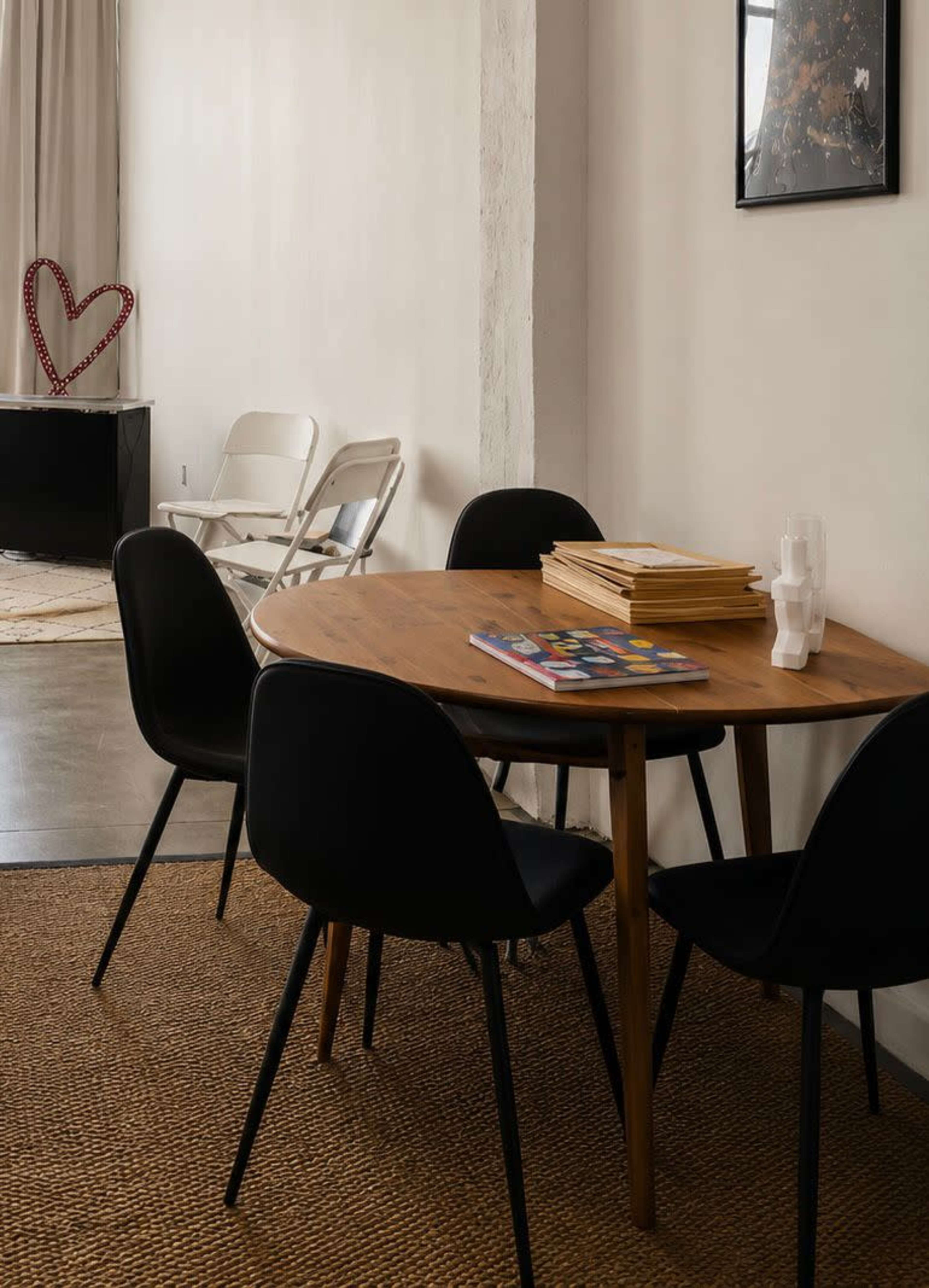 A wooden table with black chairs is set against a wall, accompanied by a stack of papers and a few plastic cups, while a heart-shaped decoration is visible in the background.