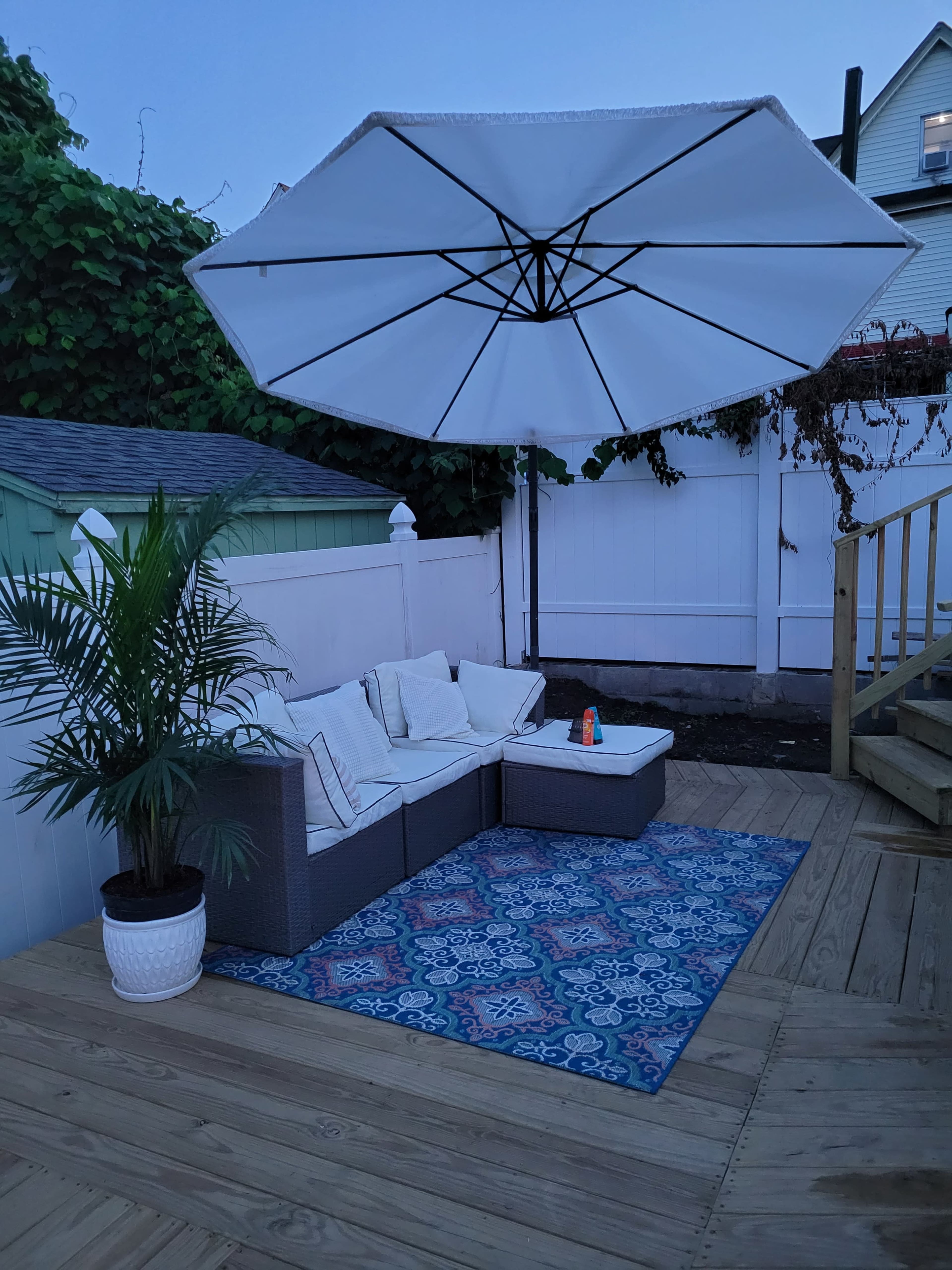 A covered seating area with a large umbrella, a sectional sofa, a small table, and a decorative rug is situated on a wooden deck surrounded by greenery.