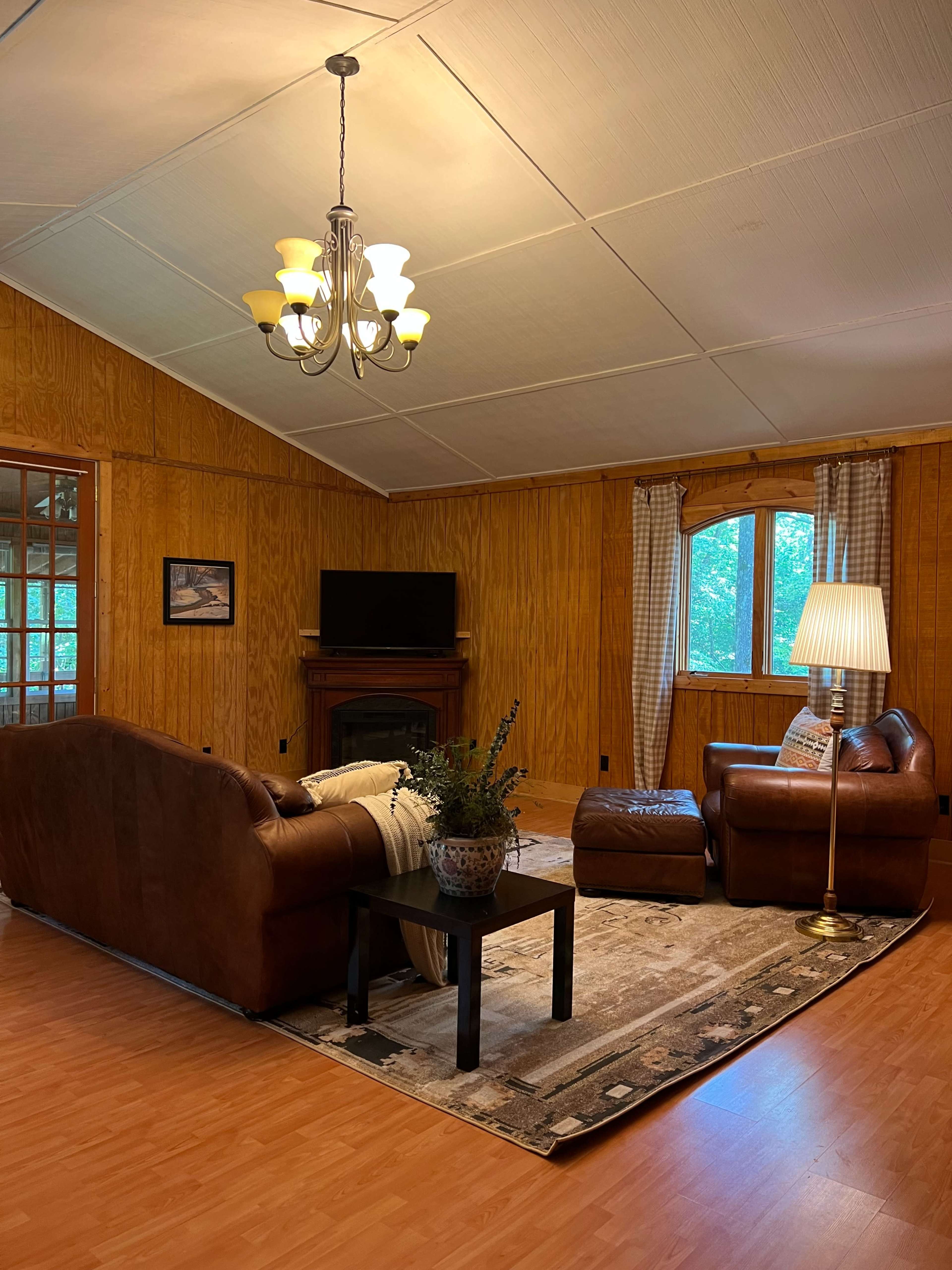 The image shows a cozy living room with brown leather sofas, a dark coffee table, a lamp, and wooden paneling on the walls.