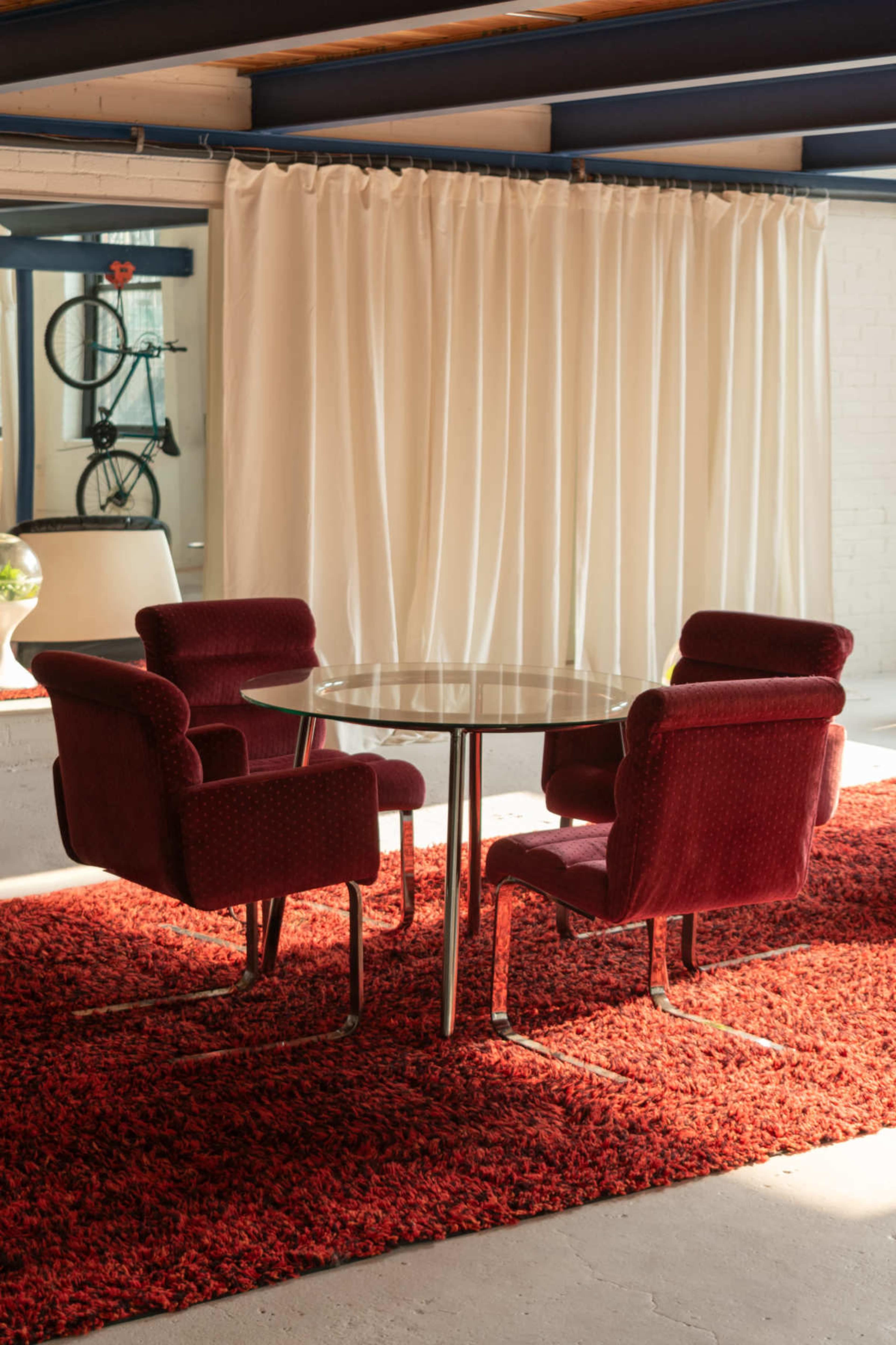 A round glass dining table is surrounded by four red upholstered chairs on a textured red rug, with a white curtain in the background.