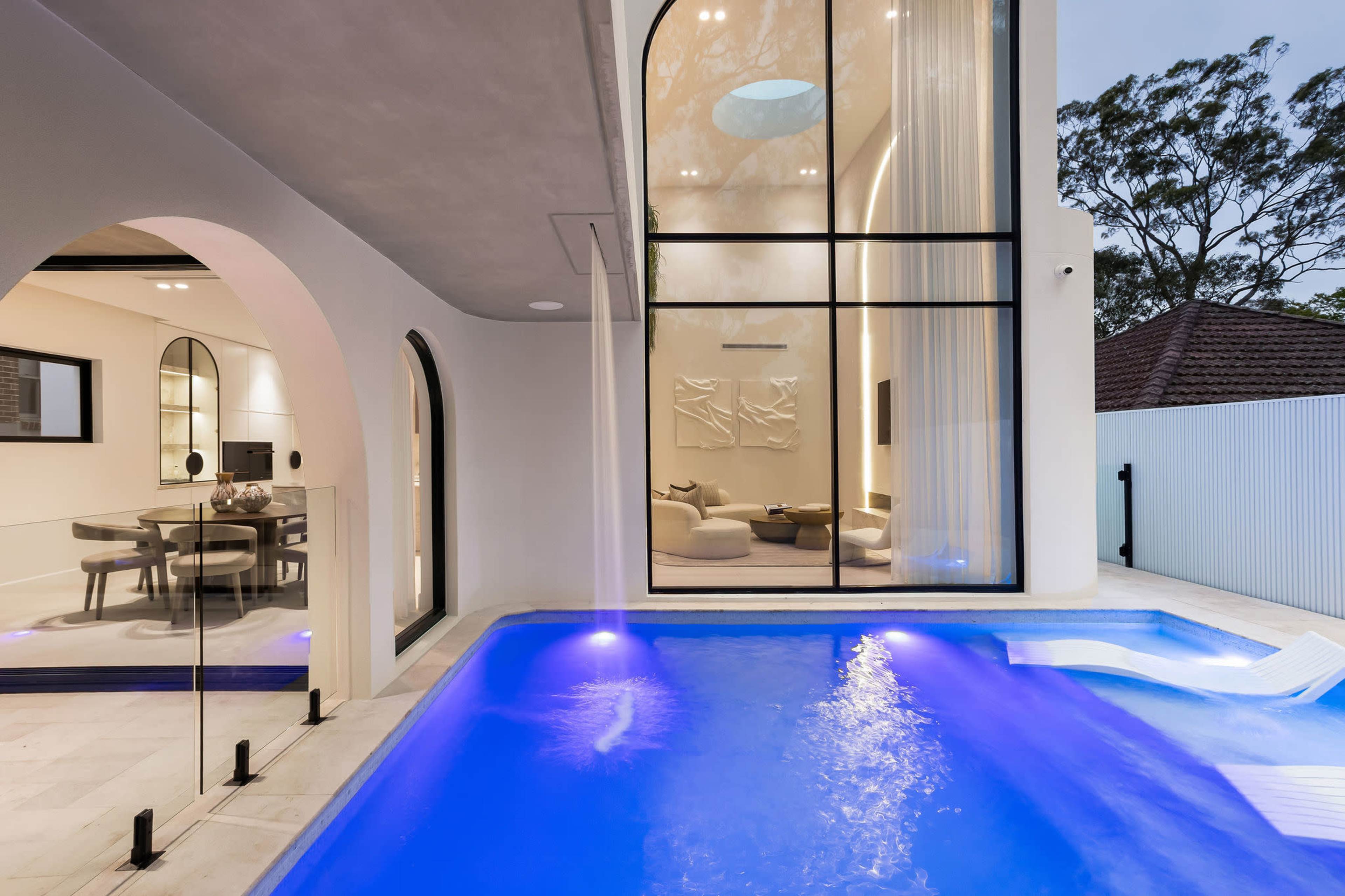 The image shows a modern villa with a clear blue pool, featuring large glass windows that reveal an elegantly designed interior space.