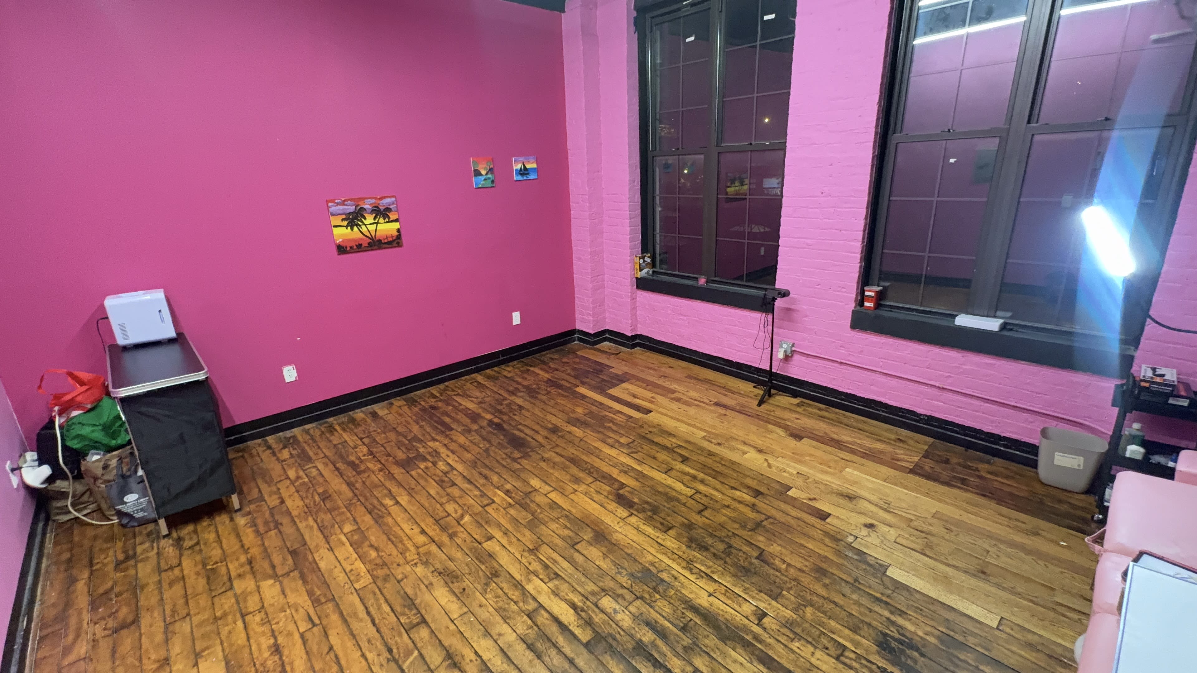The image shows an empty room with pink walls, wooden flooring, and a few pieces of furniture against the walls.