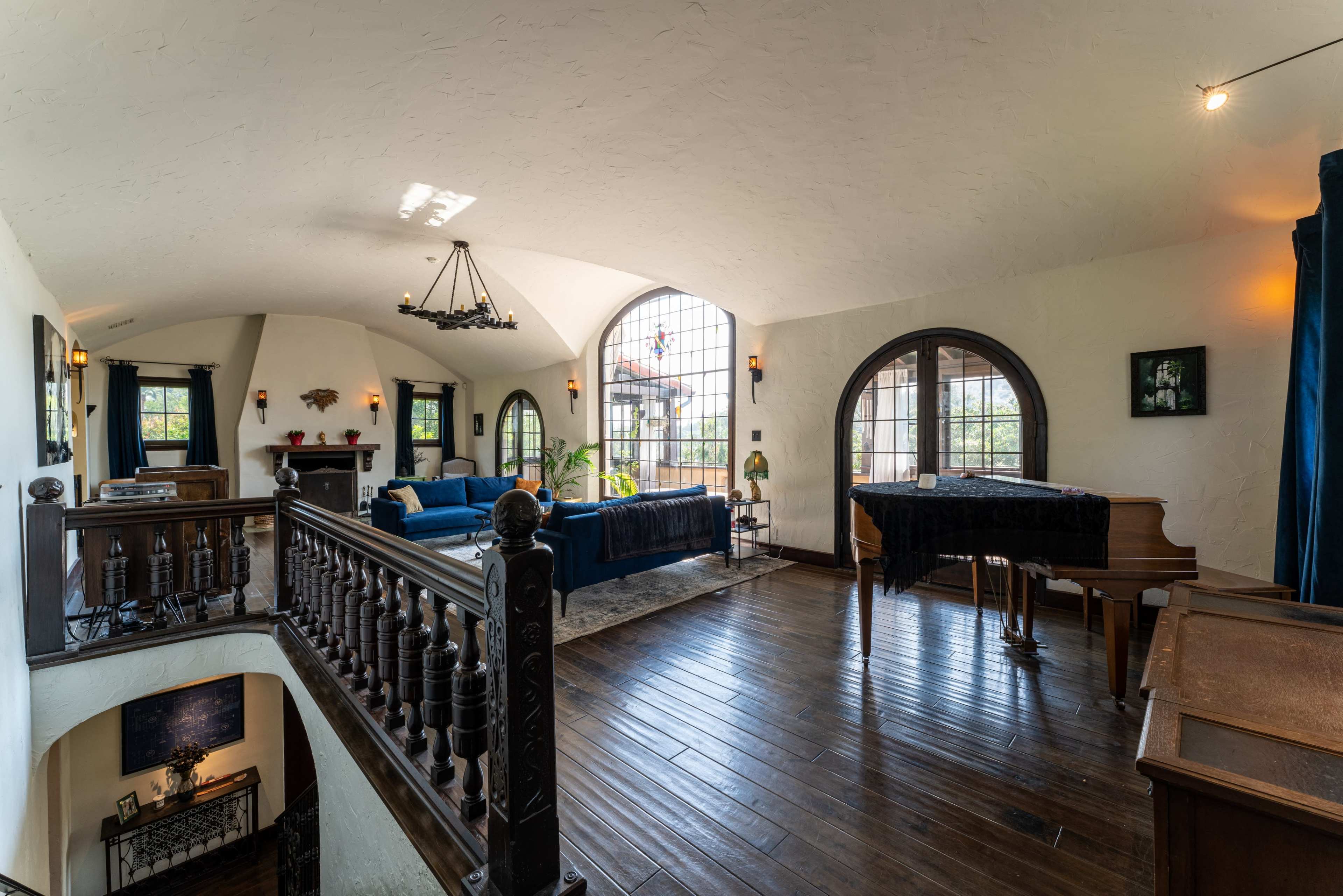 The image shows a spacious, light-filled living room featuring a grand piano, large arched windows, and a wooden balcony railing overlooking a lower level.