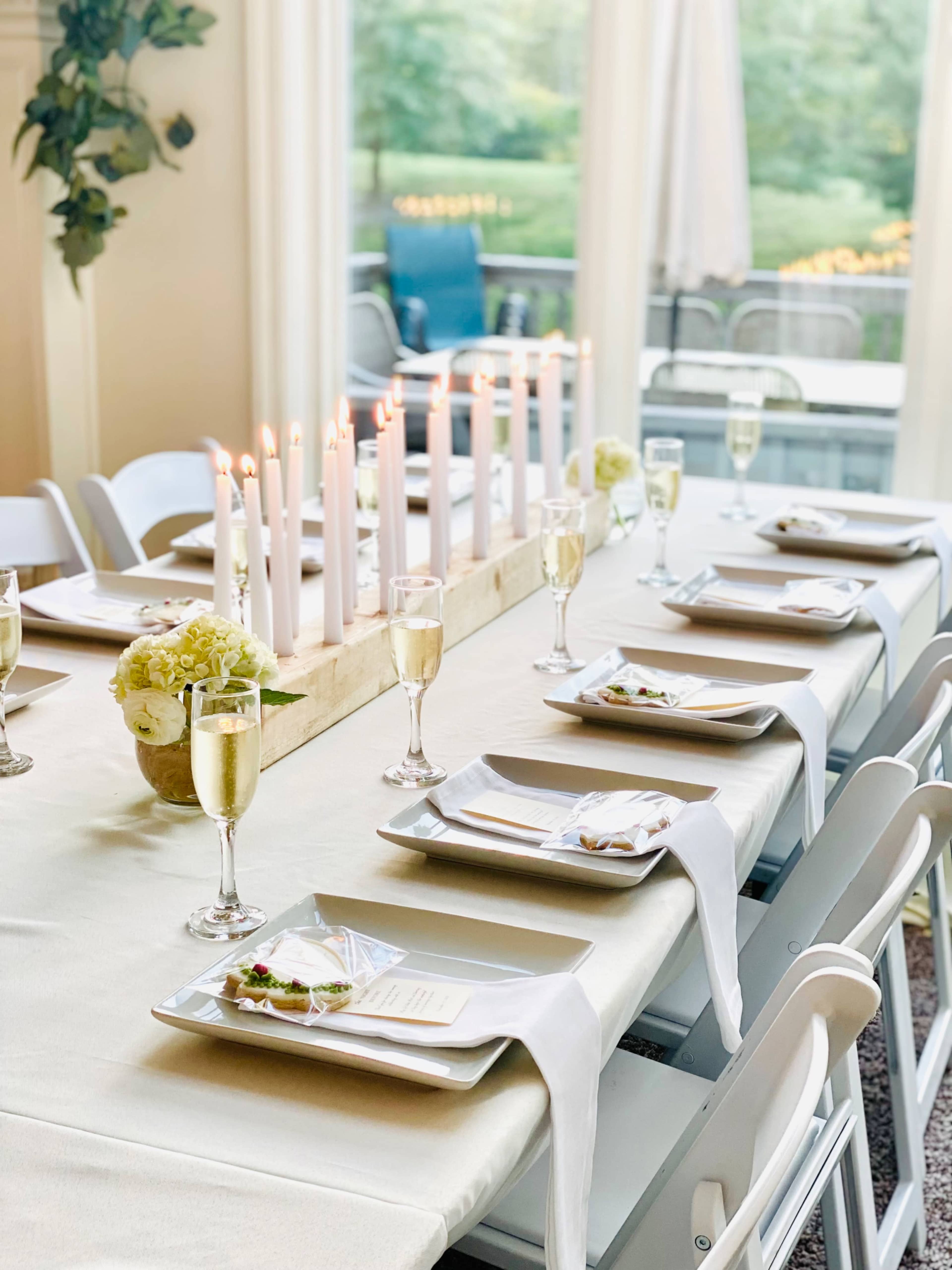 A long dining table is set with white chairs, neatly arranged plates, glasses of champagne, and a centerpiece of candles and flowers.