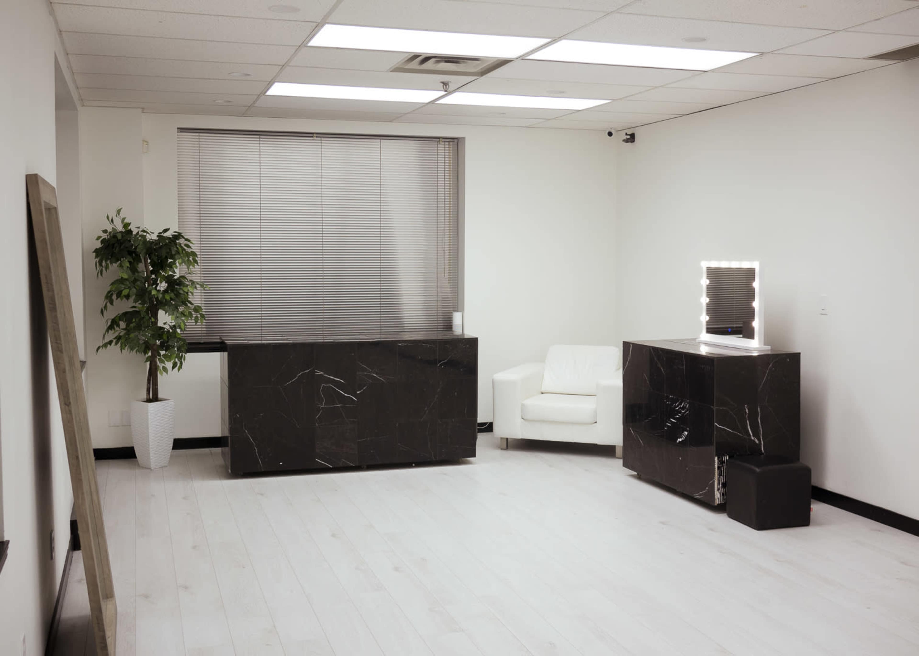A minimalist room with a white floor, a large window with blinds, a potted plant, a black marble counter, a white armchair, and a backlit mirror on a second black marble counter.