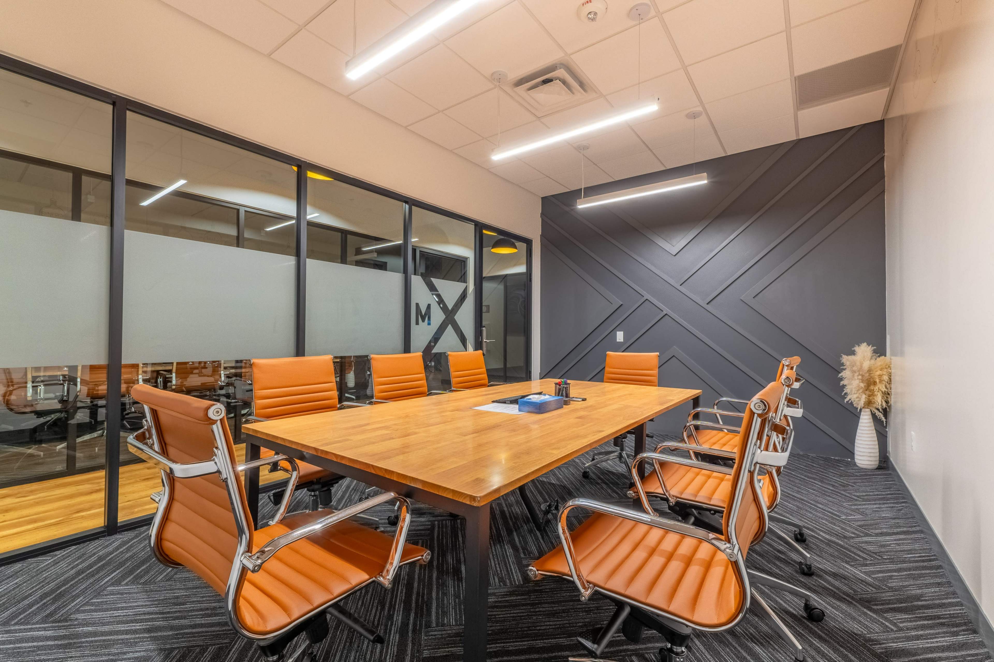 A modern conference room features a wooden table surrounded by orange leather chairs and large windows.