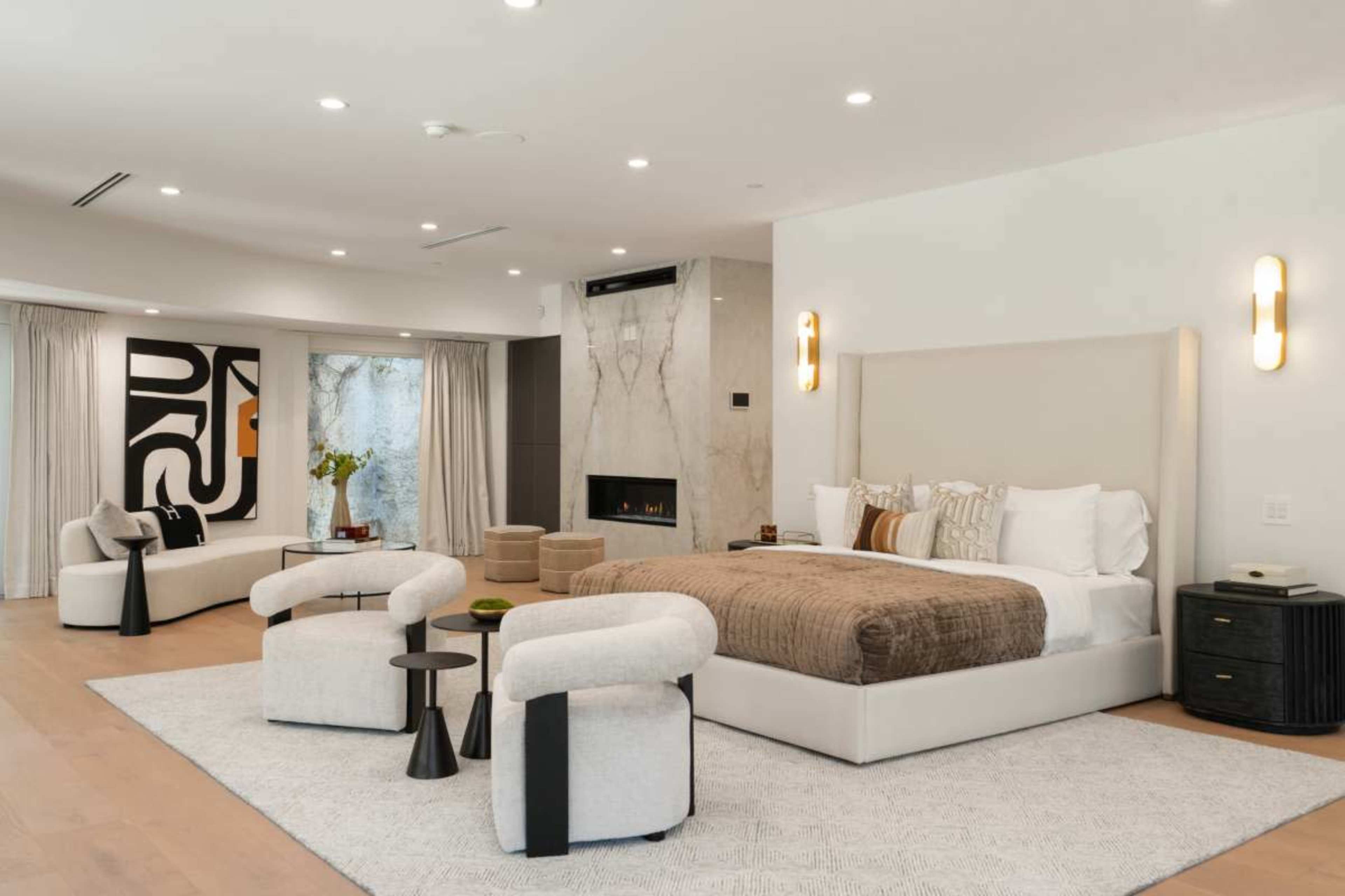 The image depicts a modern bedroom featuring a large bed with a neutral-colored headboard, two stylish chairs, and a minimalist decor aesthetic.