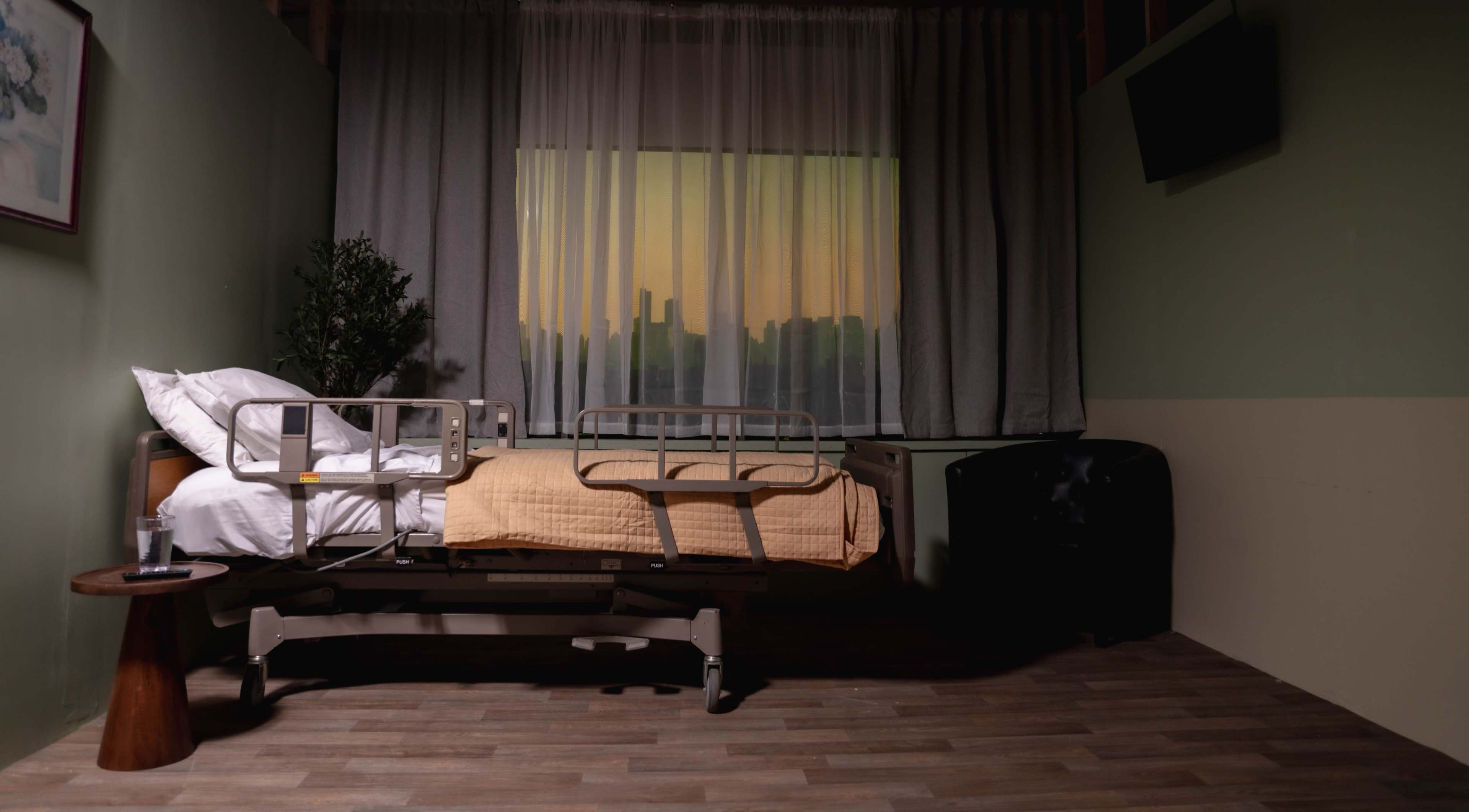 The image shows a hospital room with an unoccupied bed next to a window covered by sheer curtains, revealing a city skyline at sunset.