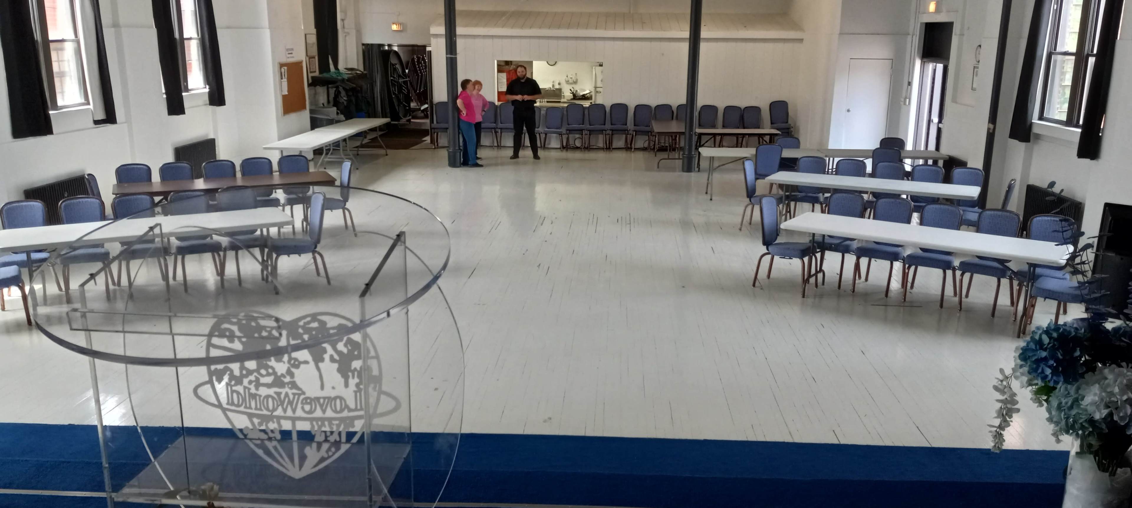 The image shows a spacious, empty room set up with rows of tables and chairs, featuring a small stage area with a transparent podium in the foreground.