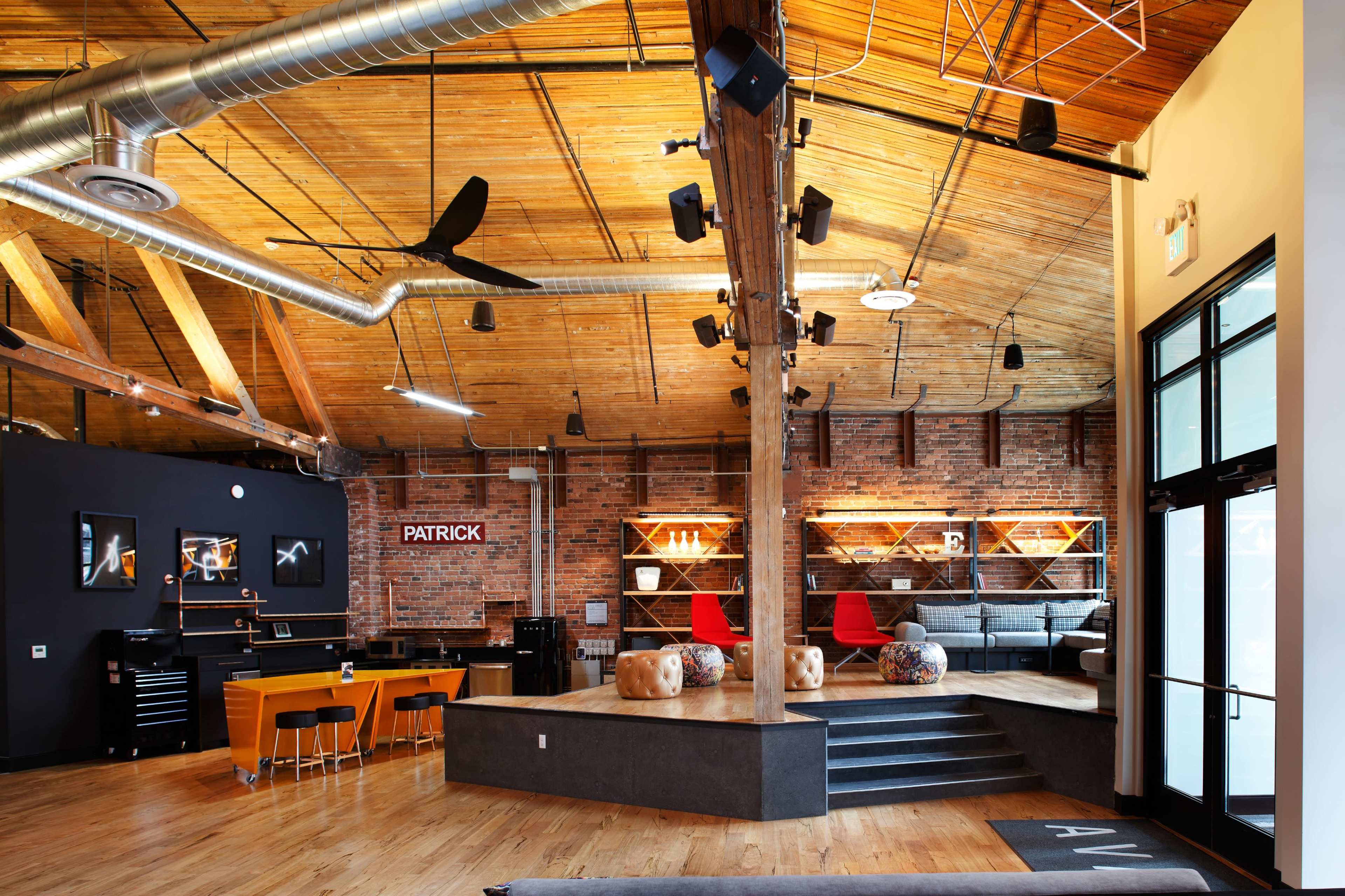 A spacious, industrial-style interior with wooden beams, exposed brick walls, a central seating area, and an elevated platform surrounded by modern furniture.