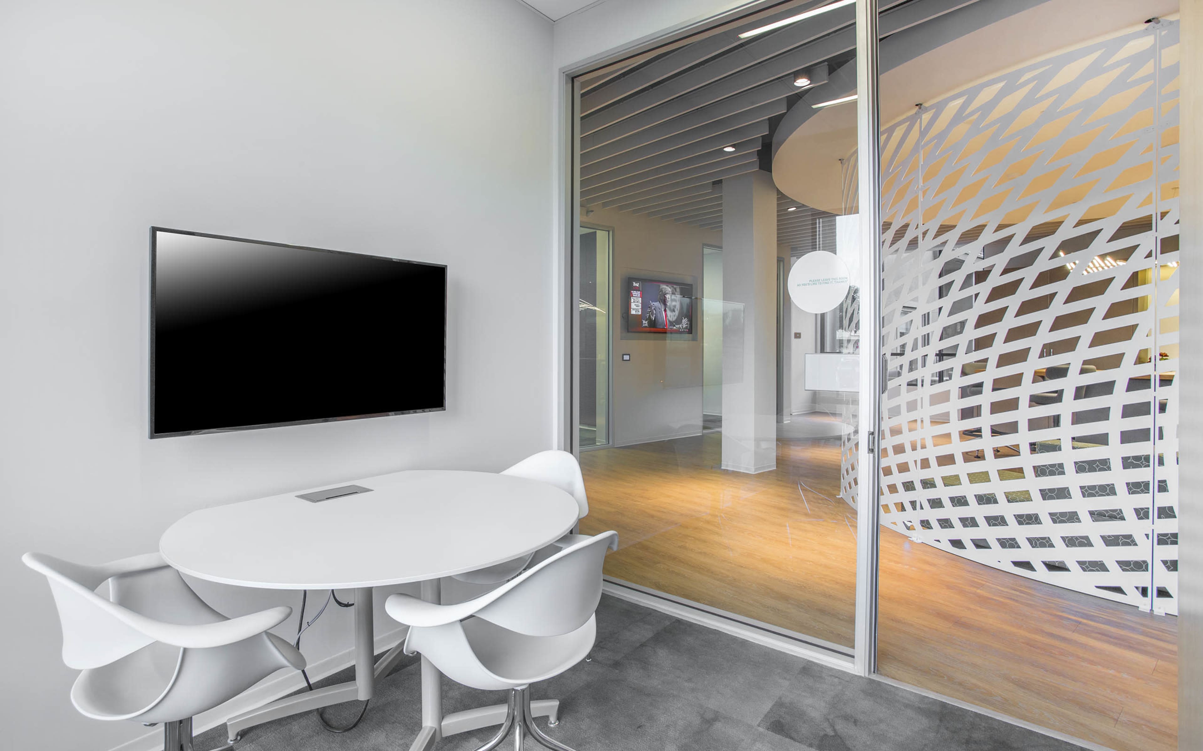 A conference room features a round table with white chairs and a wall-mounted television, along with glass walls that provide a view of a connected space.