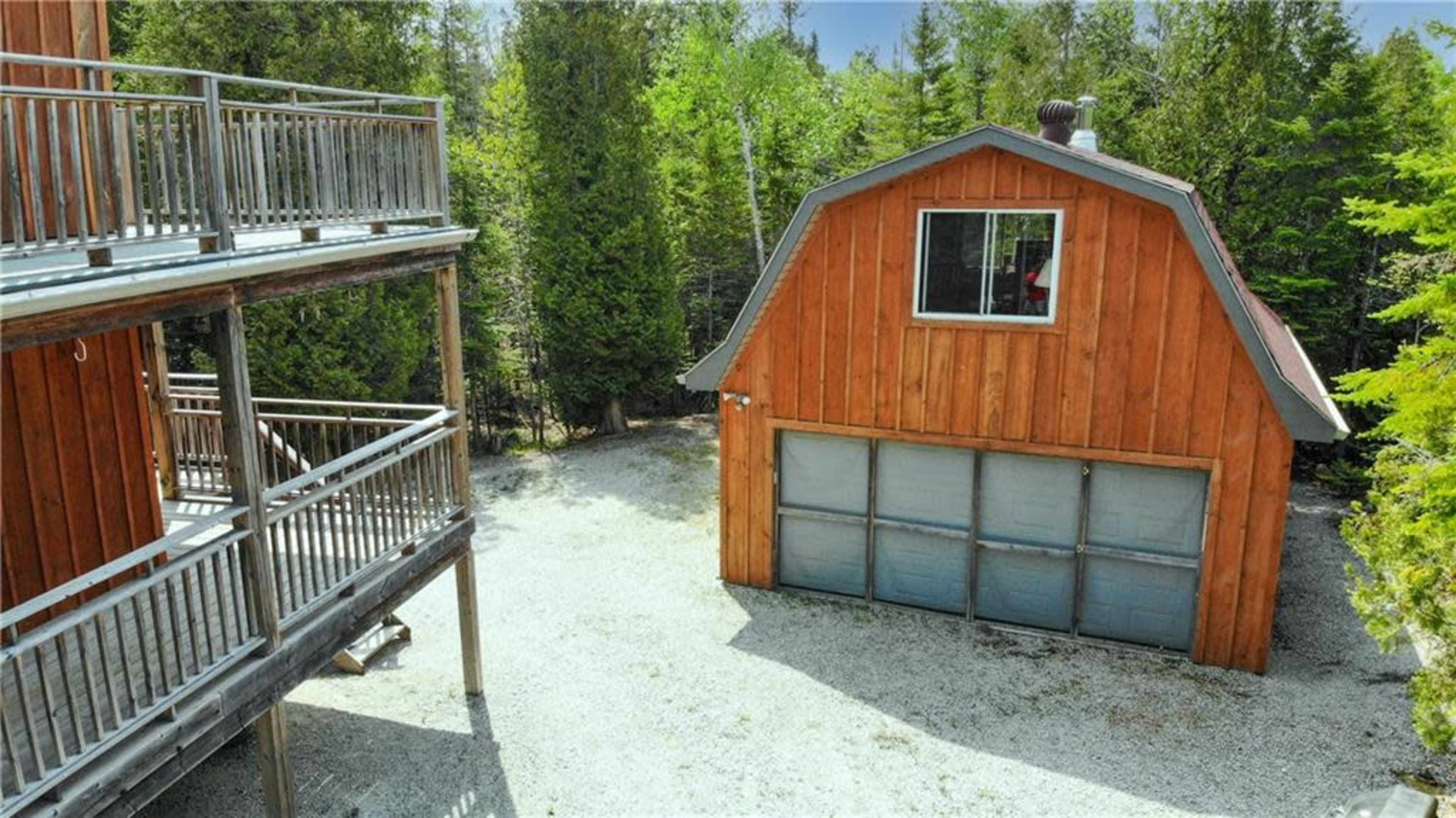 A wooden structure with a gray garage door stands next to a two-story building with a deck, surrounded by greenery.