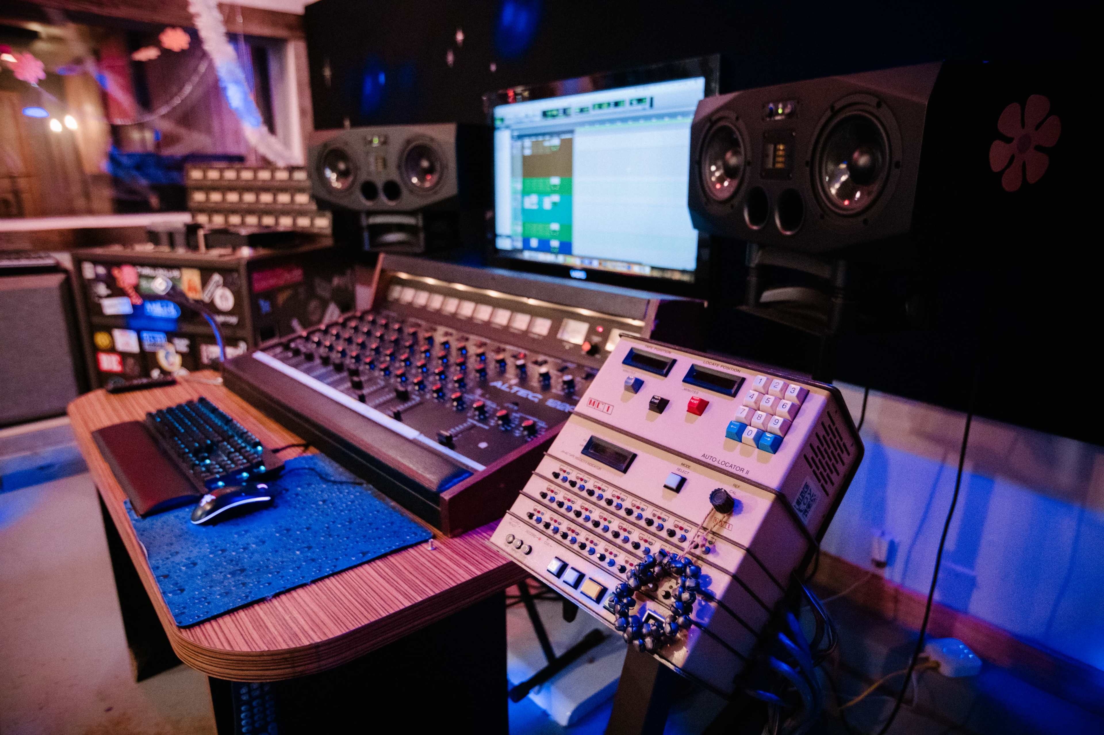 A music production workstation featuring a mixing console, studio monitors, a computer screen displaying digital audio software, and various music production equipment.
