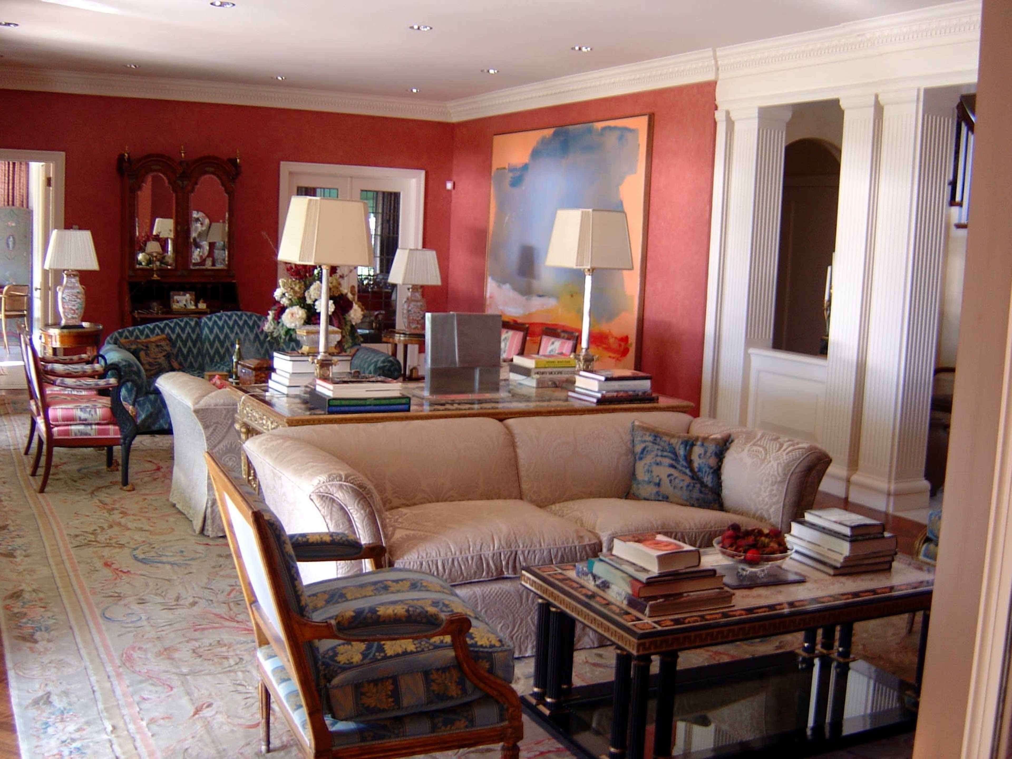 The image shows a spacious living room with beige sofas, decorative tables stacked with books, and colorful artworks on the walls.