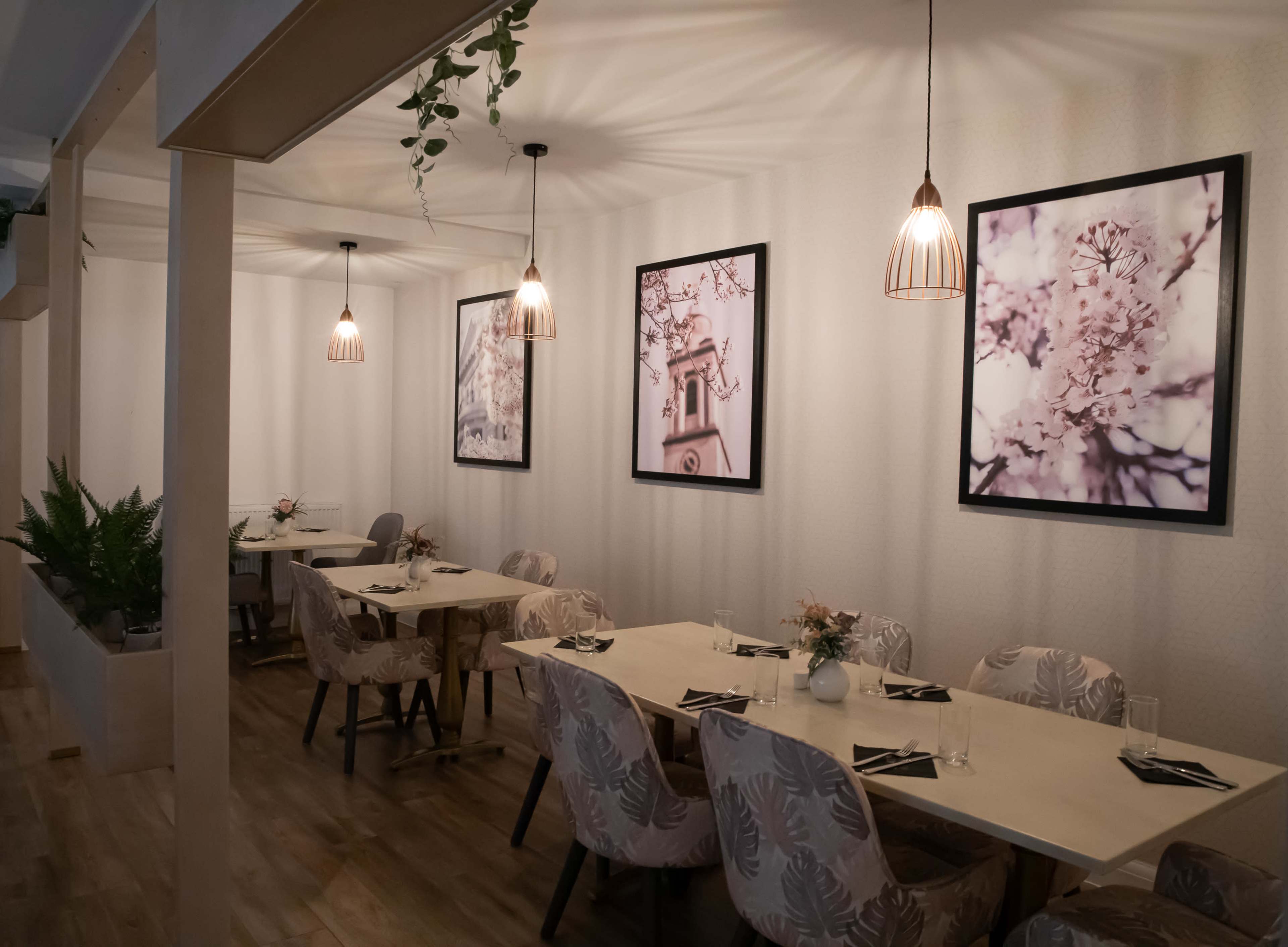 A modern dining area with tables set for multiple guests, adorned with framed floral prints on the walls and pendant lights overhead.