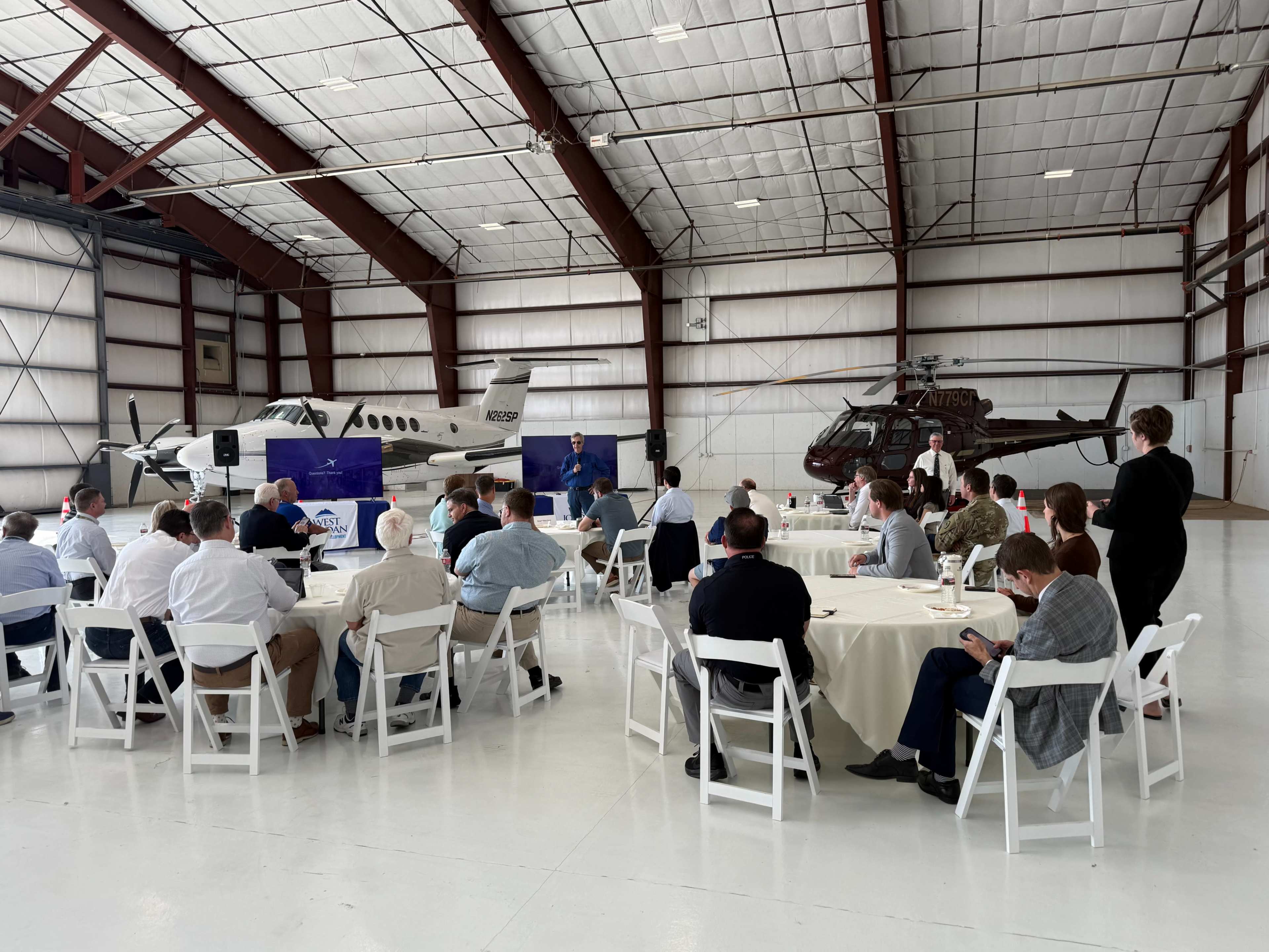 A group of people is seated at round tables in a hangar, with an airplane and a helicopter visible in the background.